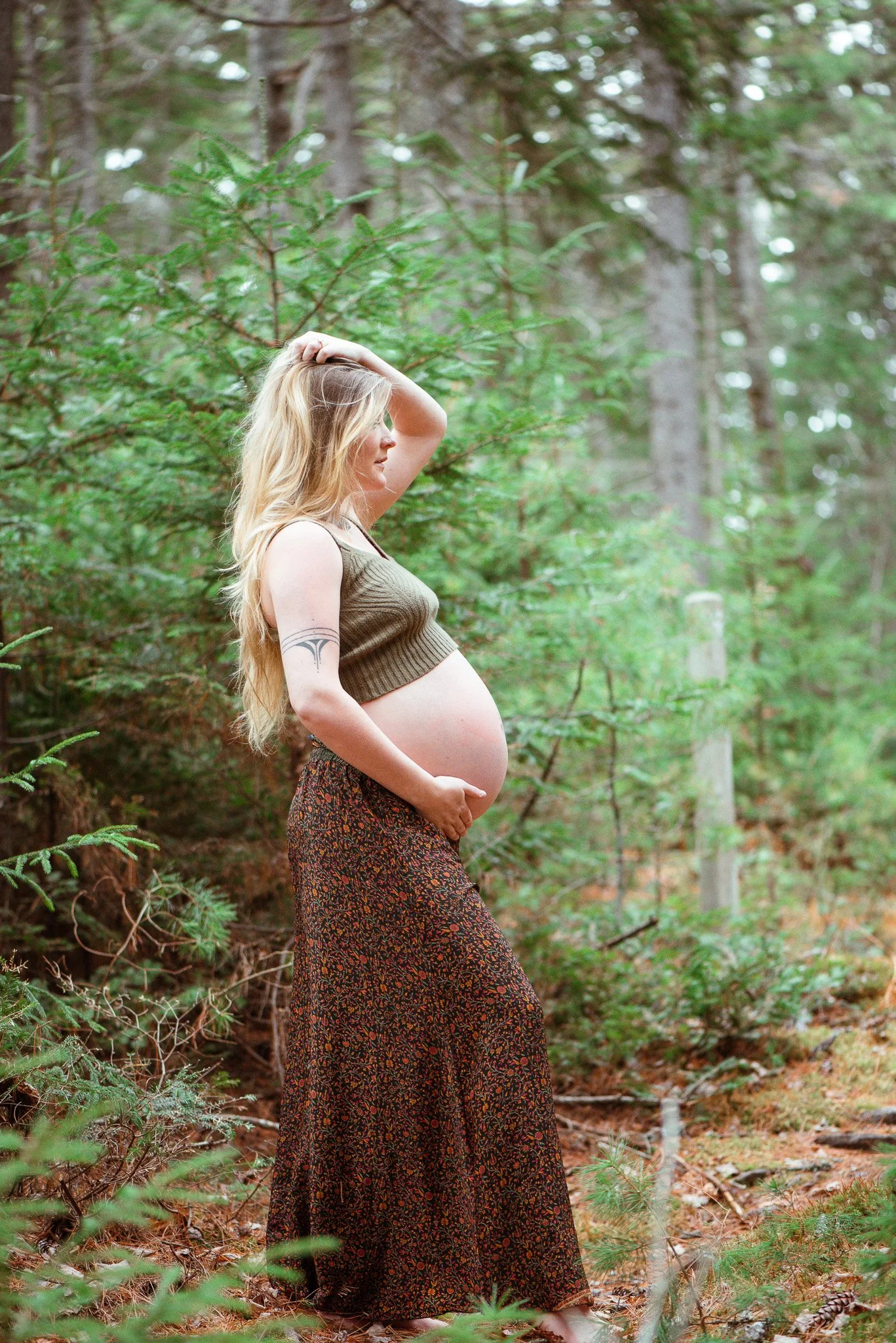 A pregnant woman with long blonde hair standing in a forest with green trees and pine needles on the ground, wearing a sleeveless brown top and a long patterned skirt, looking to the side with one hand on her head.