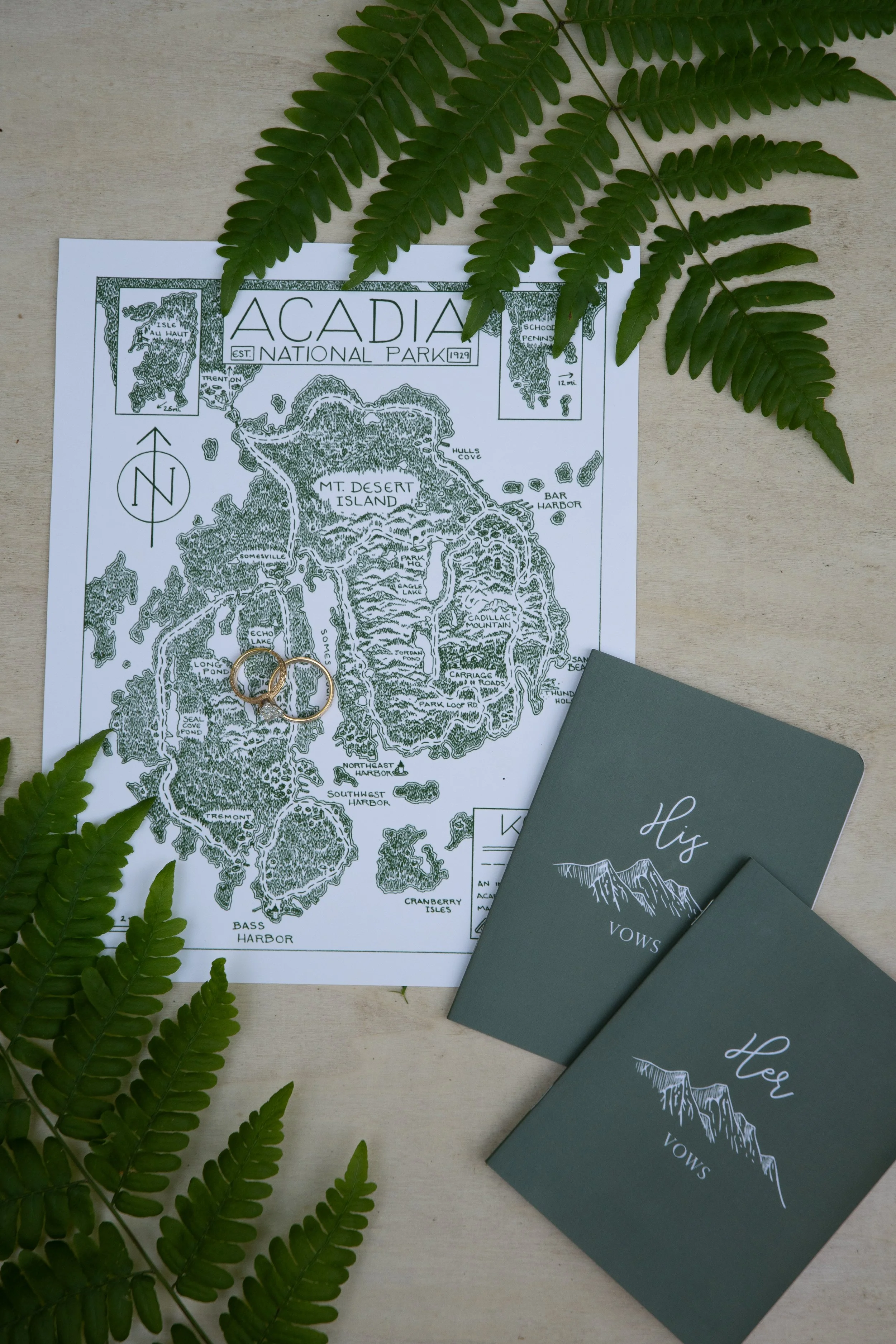 A top view of a map of Acadia National Park with two wedding rings resting on it, surrounded by green fern leaves and two vow booklets labeled 'His Vows' and 'Her Vows'.