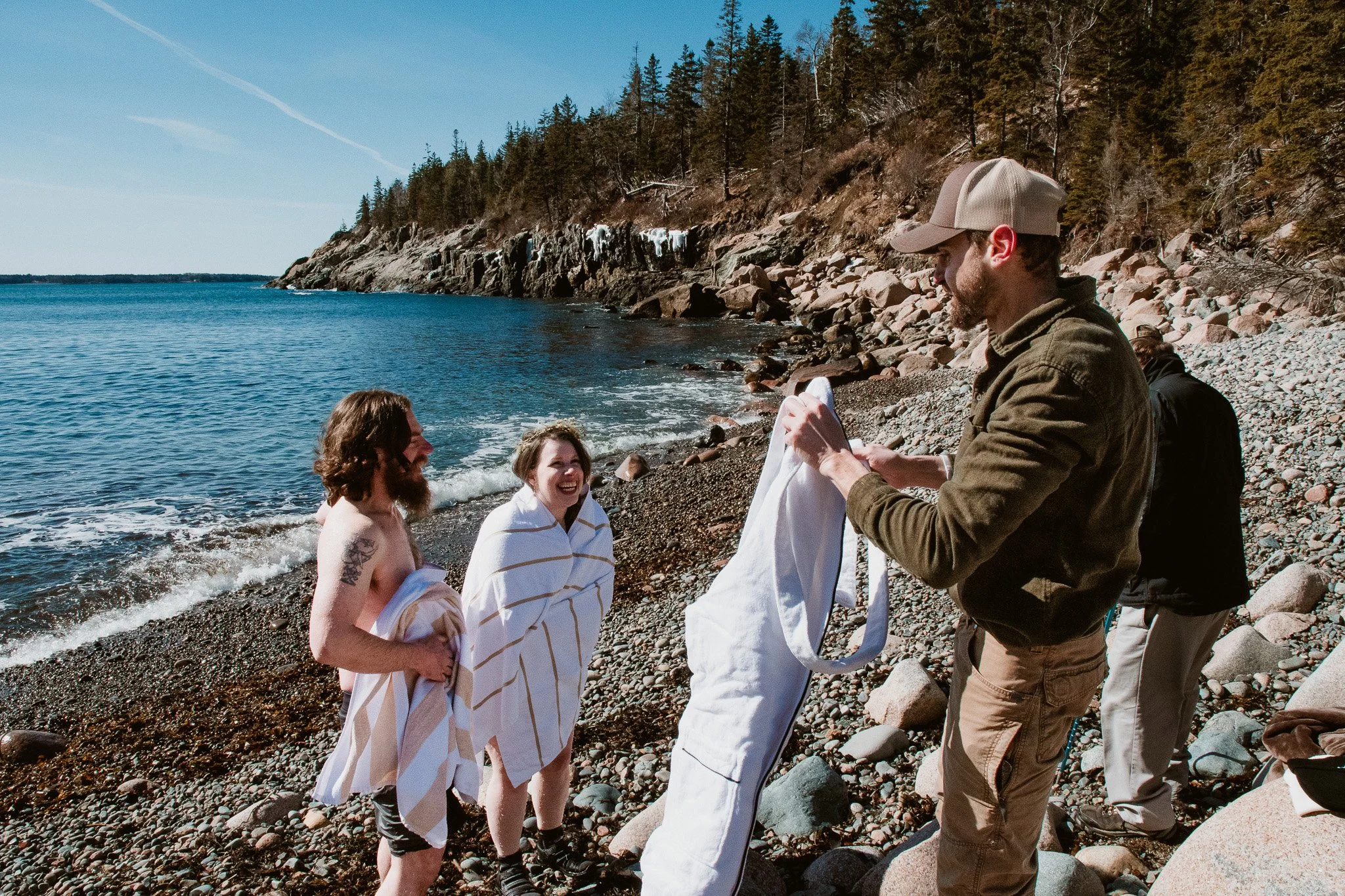 People on a rocky beach preparing for a dunking, with two women already wet and smiling, and a man holding a white towel, in a scenic outdoor setting with ocean and trees.