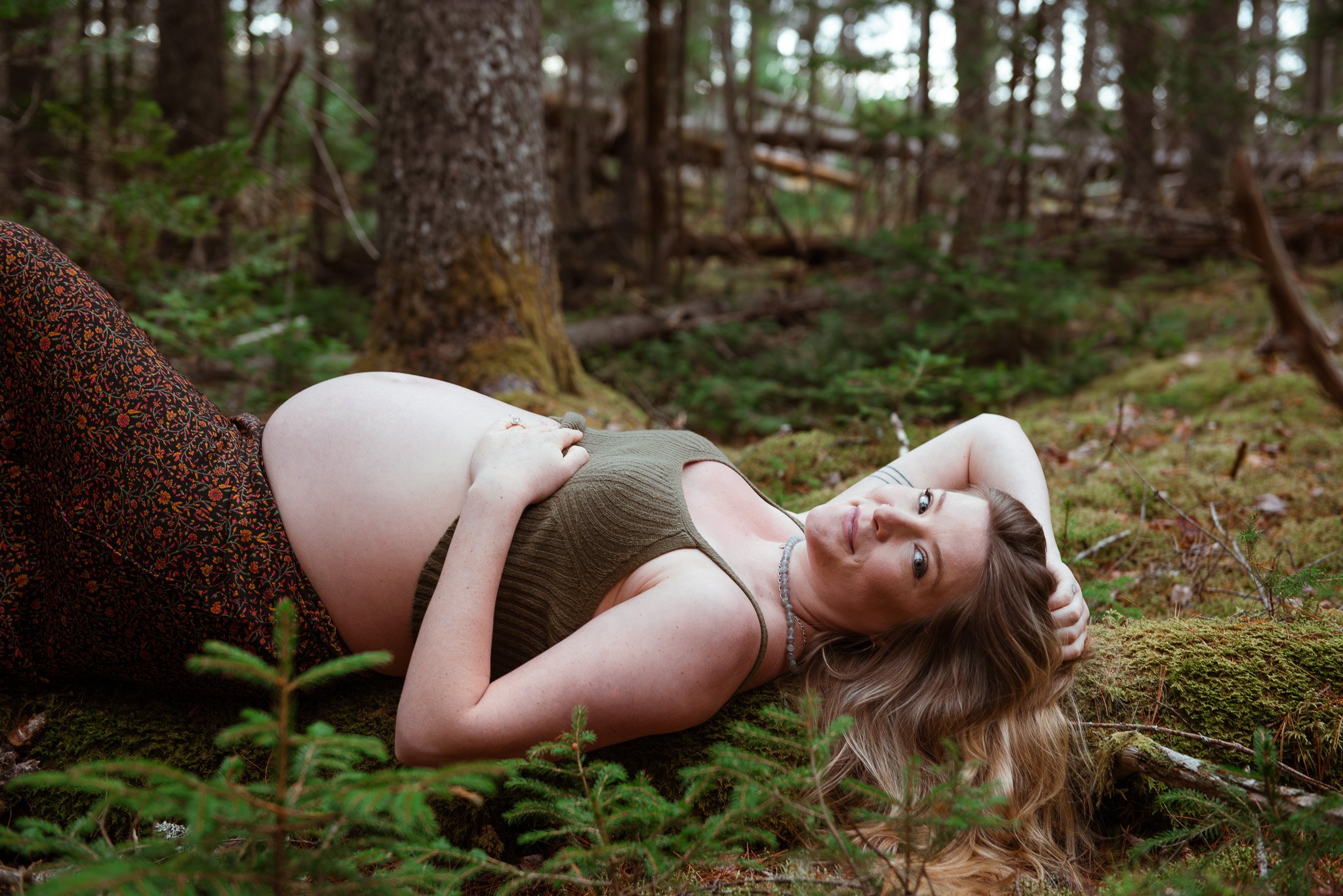 A woman with long hair lying on her back on forest ground, wearing a brown top and patterned skirt, looking at the camera with her head resting on her arm, surrounded by trees and greenery.