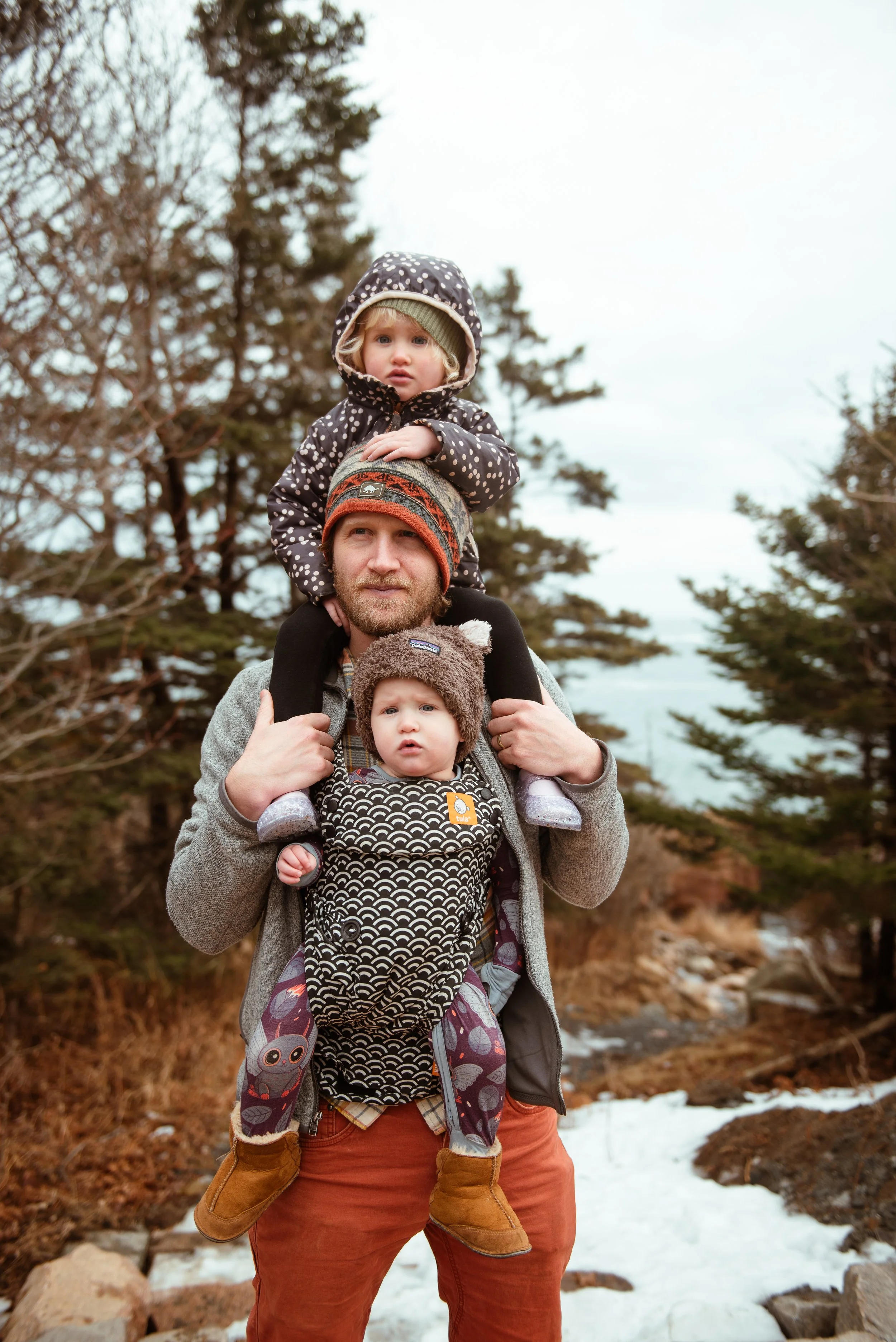 A man carrying two small children on his shoulders outdoors near trees and snow.