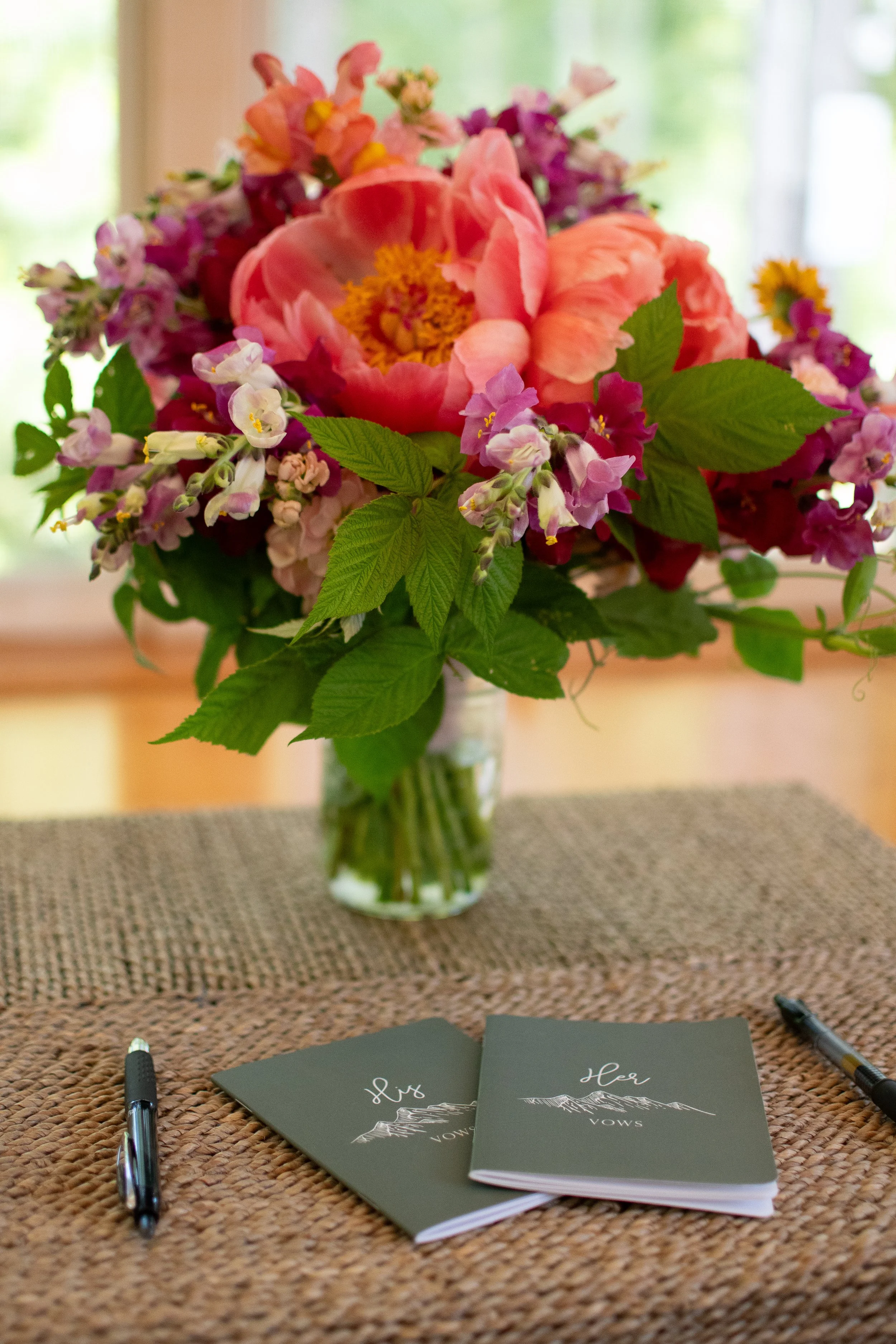 A flower bouquet with pink, purple, and yellow flowers in a glass vase on a table, with two black pens and vow booklets that say "Her Vows" placed in front of the bouquet.