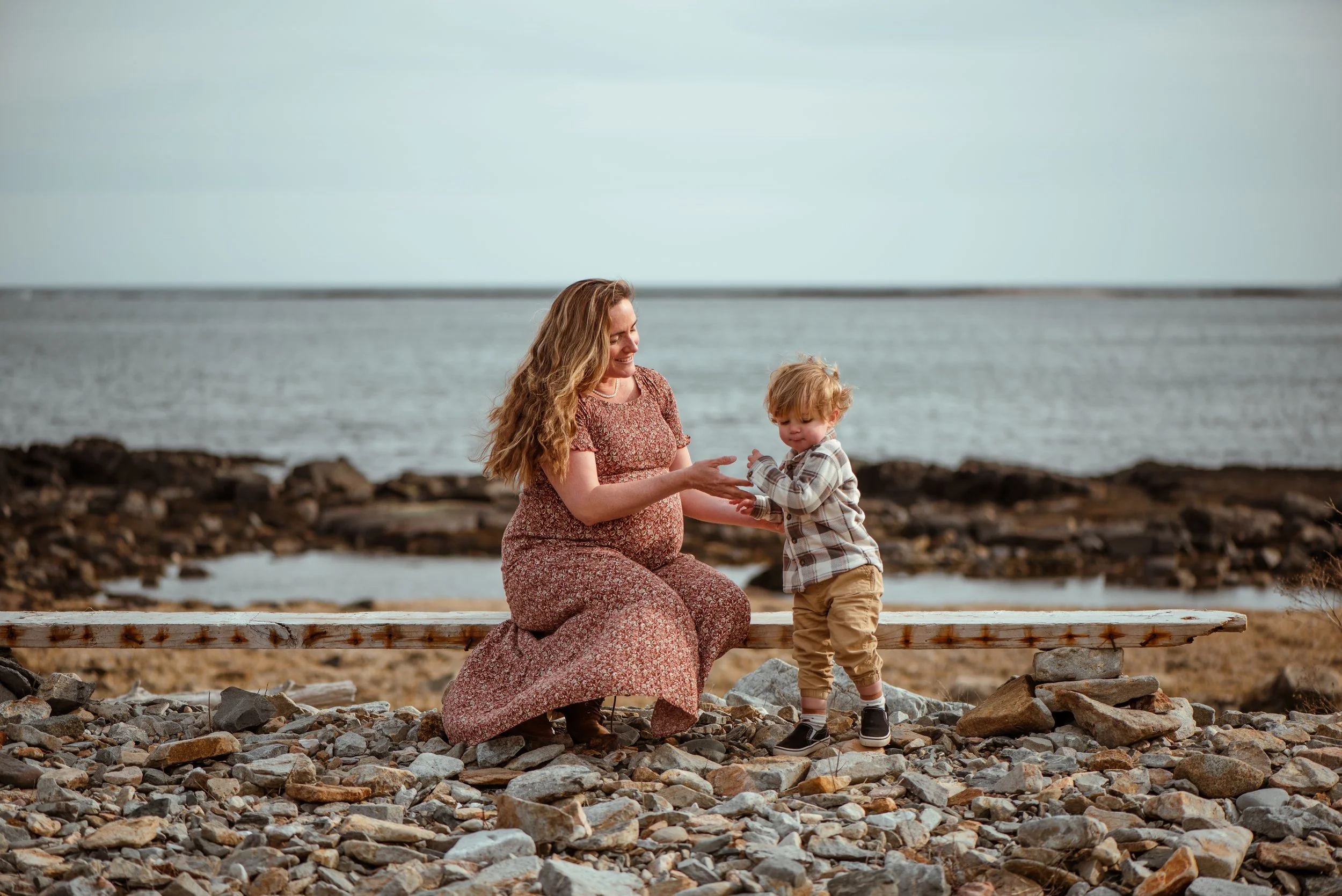 A pregnant woman in a long floral dress kneeling on a rocky beach, smiling as she interacts with her son in a plaid shirt and khakis, with the ocean in the background.