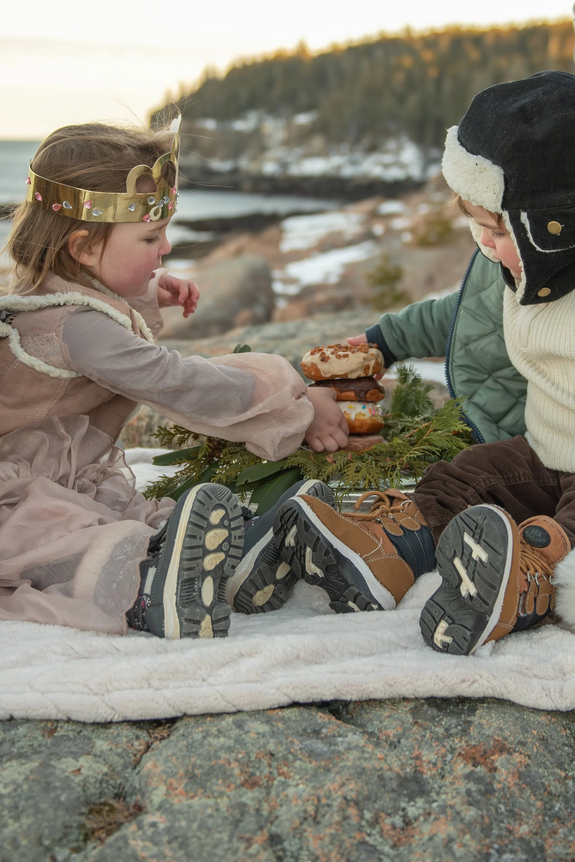 Two children dressed in winter clothing, sitting on a white blanket outdoors near a rocky shoreline with snow and a distant forested hill. The girl wears a crown and the boy wears a warm hat, and they are sharing a large frosted donut.