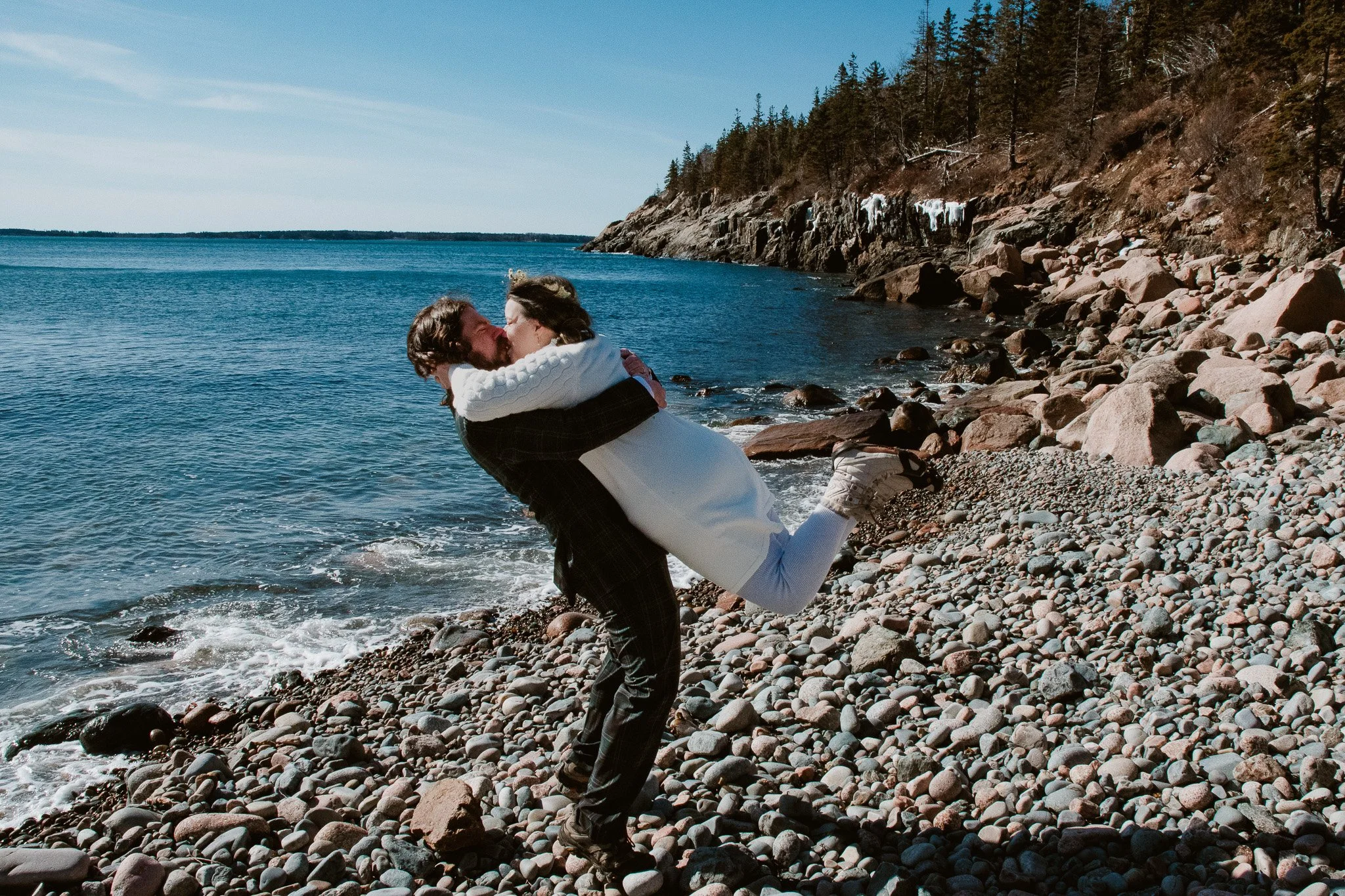 A man in dark clothing lifts a woman in white onto a rocky beach near the water, with a cliff and trees in the background, on a sunny day.