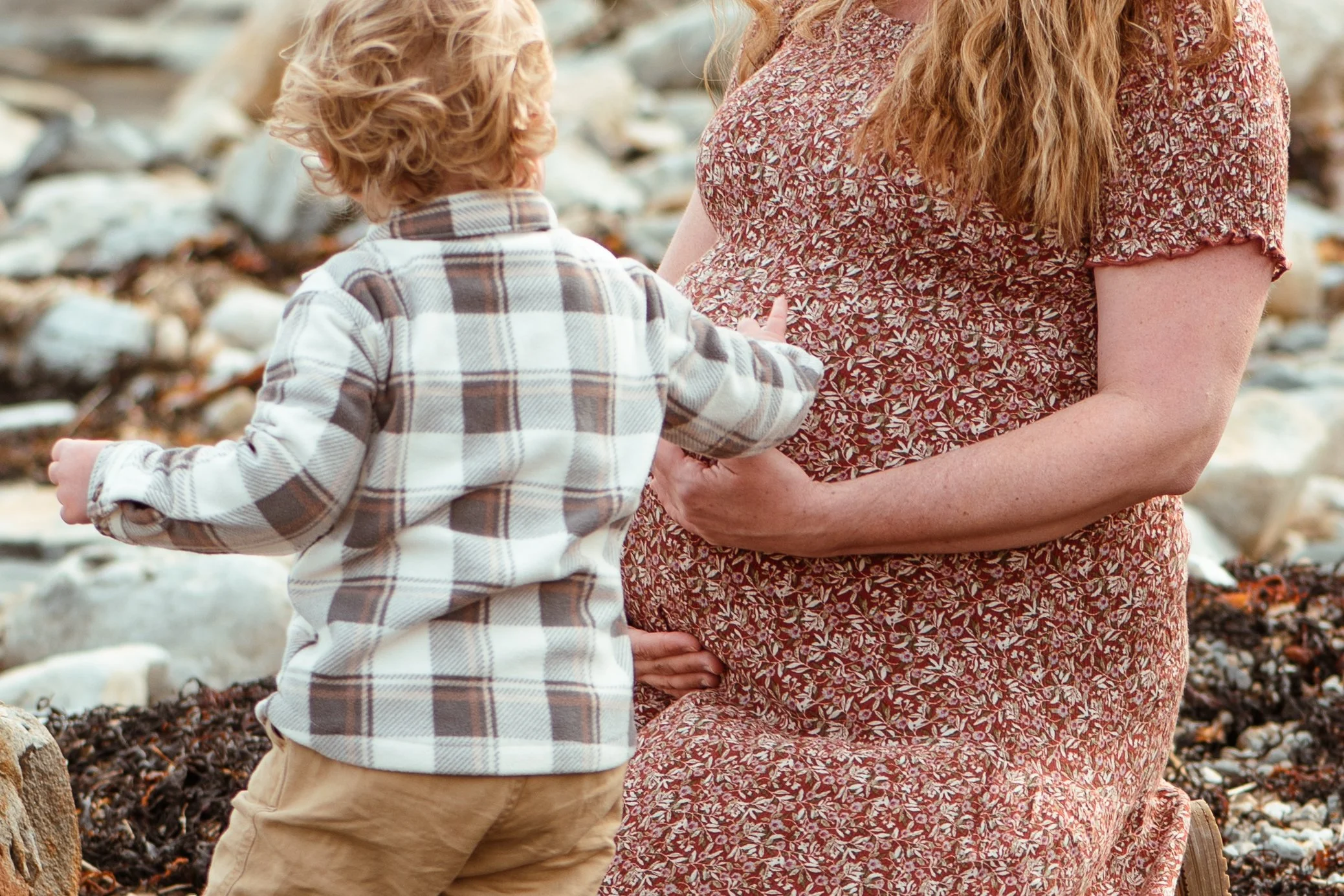A pregnant woman and her son standing outdoors on a rocky beach. The boy is wearing a plaid shirt and khaki pants, while the woman is dressed in a floral dress. The woman is touching the boy's arm, and they are engaged in a moment of connection.