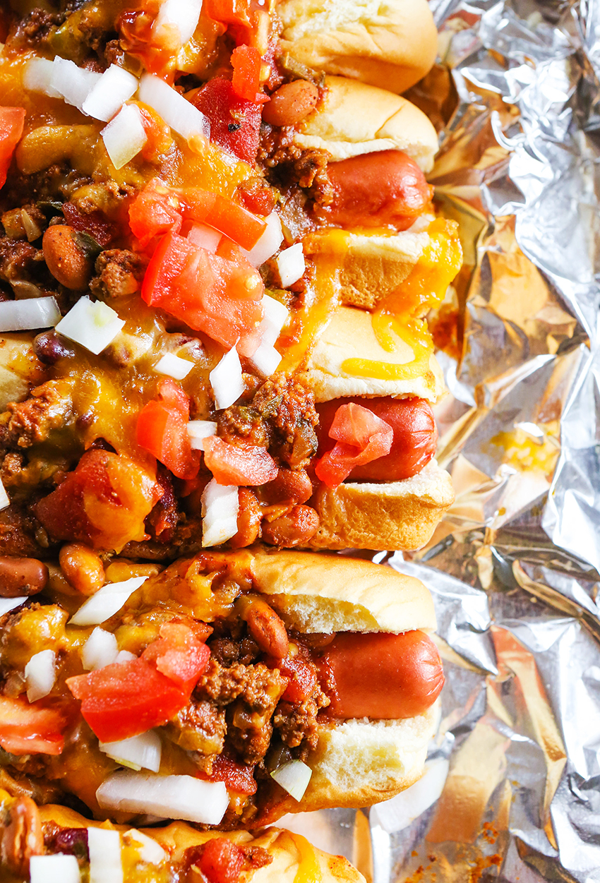 Close up vertical shot of chili dogs lined up on aluminum foil.