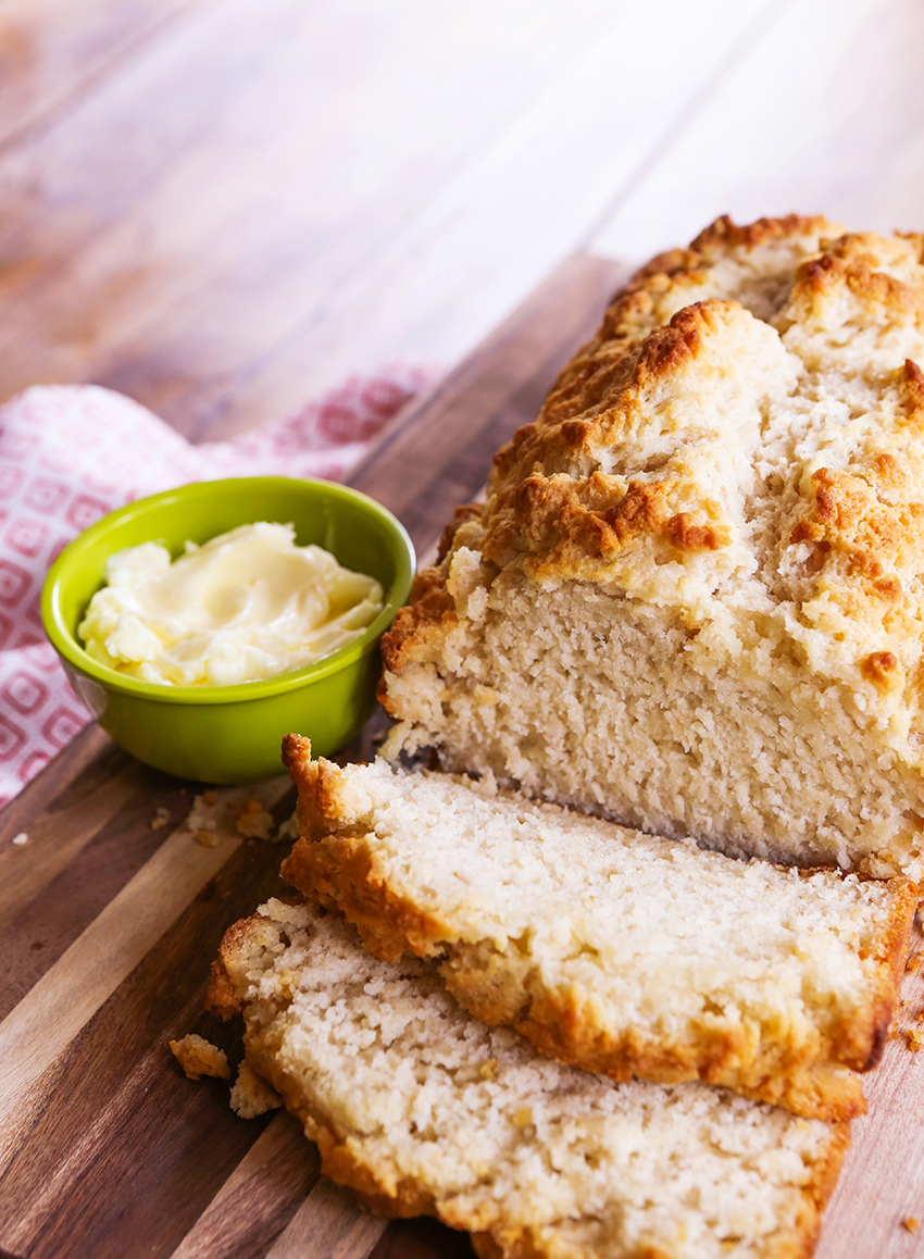 A small cup of softened butter next to a loaf of best beer bread with two slices cut from it