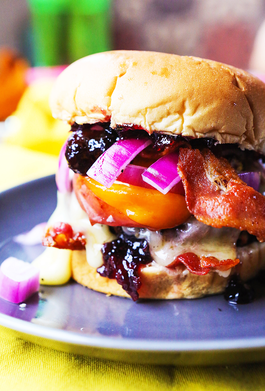 A side view of a grilled burger on a bun with grilled peaches, cherry and brie jam and bacon on a plate