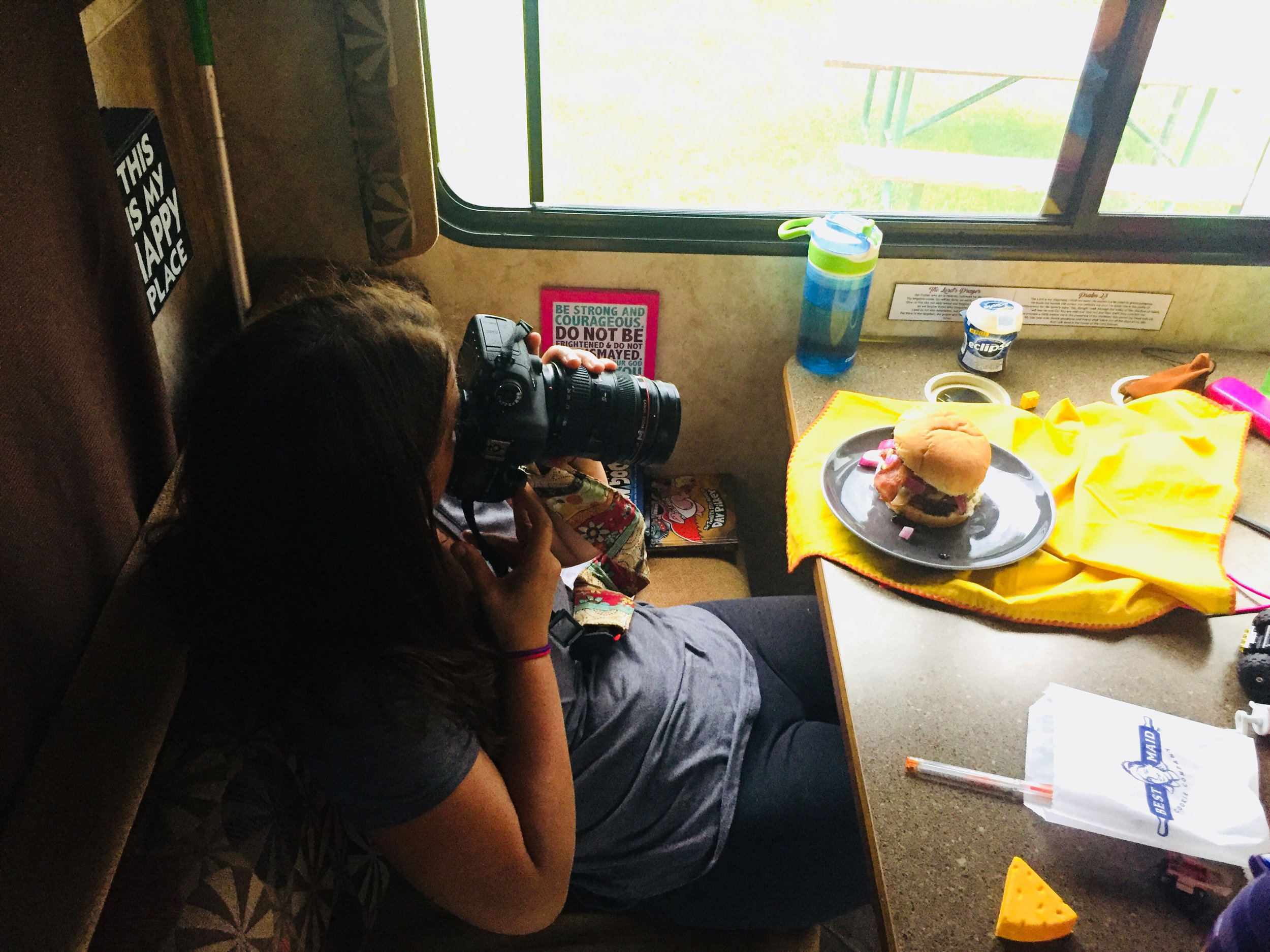 Overhead view of photographer taking a picture of a plated burger with grilled peaches, cherry and brie jam on a plate sitting on a table in an RV.