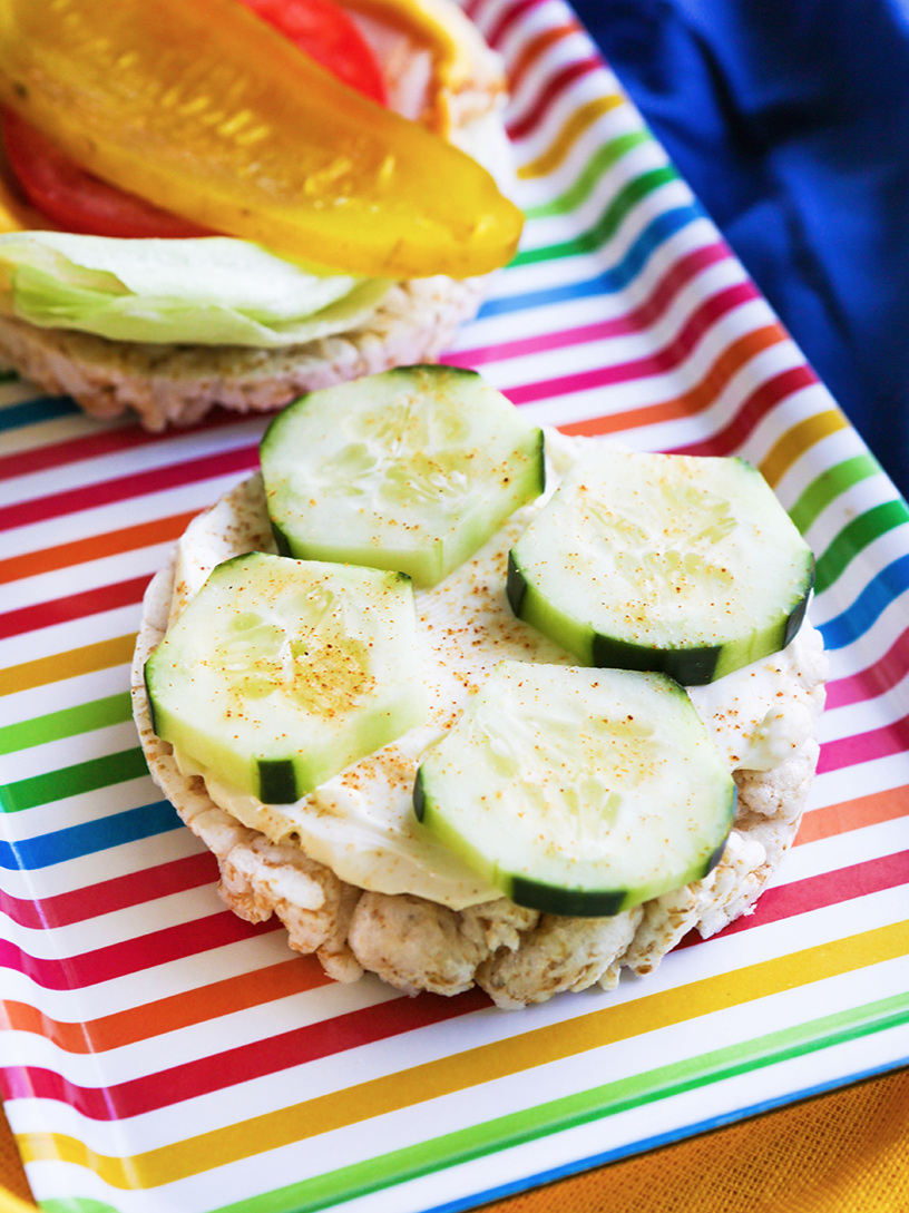Colorful striped tray displaying a rice cake that has cucumbers and cream cheese and another with turkey, pickles lettuce tomato