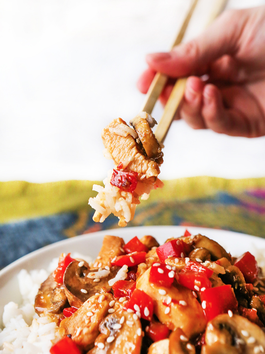 Hands holding chopsticks with a bite of rice and hoisin chicken in between over a plate of rice and hoisin chicken