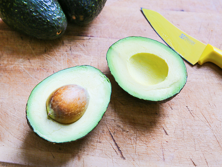 A sliced in half avocado with the seed still inside and a yellow knife sitting next to the fruit