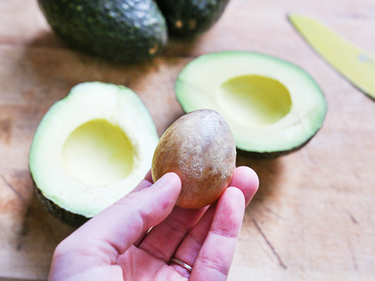 A hand hovering over a cutting board with an avocado cut open and the hand holding the seed