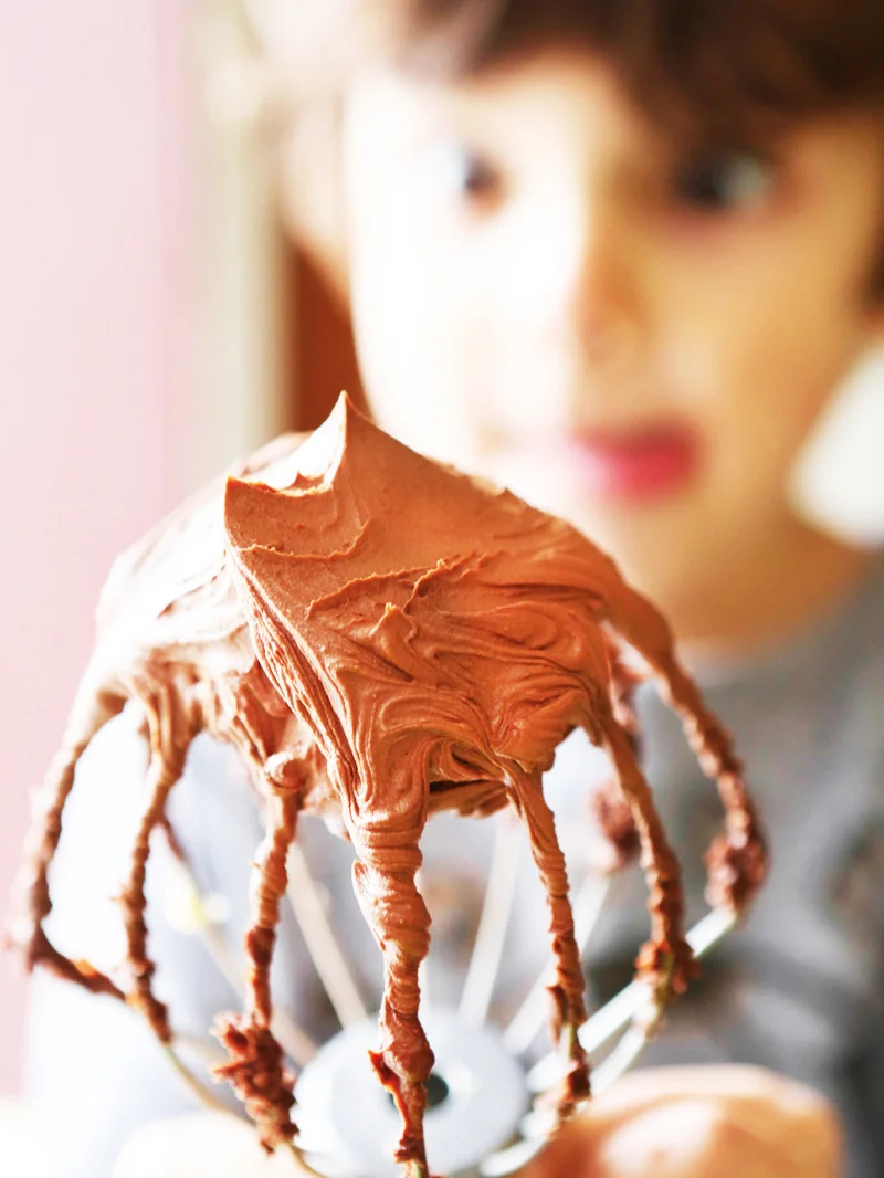 cute boy in background looking eagerly at whisk attachment filled with buttercream frosting