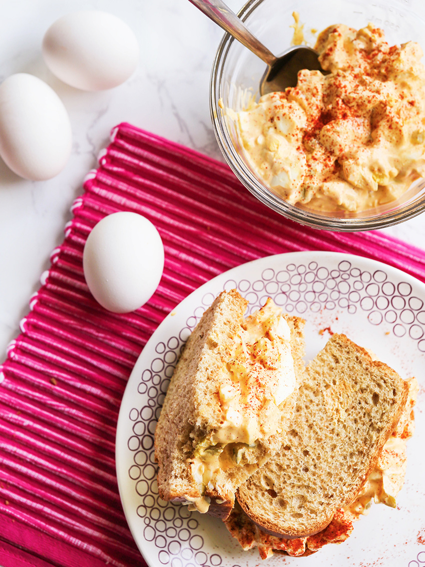 Egg salad sandwich cut in half on a plate next to eggs in the shell on the placemat next to it with a bowl of egg salad at the top of the picture