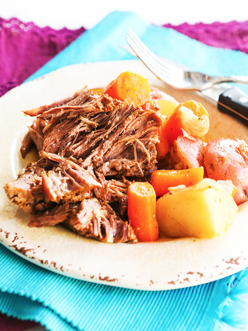 plate full of pot roast, cooked potatoes and carrots with a fork and knife on the plate. 