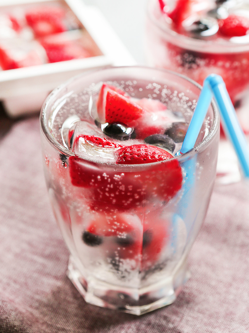 Close up of a glass full of water and fruit ice cubes with a blue straw and a ice cube tray in the background. 