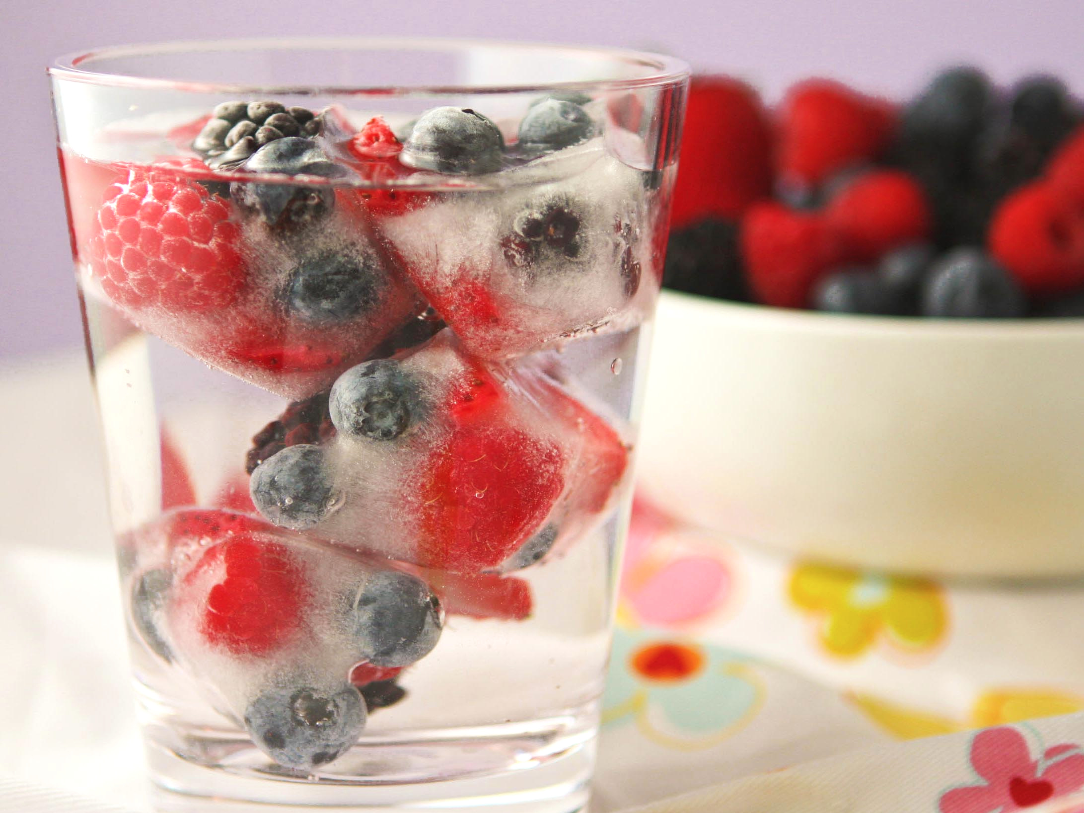 side view of a glass of water with fruit ice cubes and a bowl of fresh fruit behind it