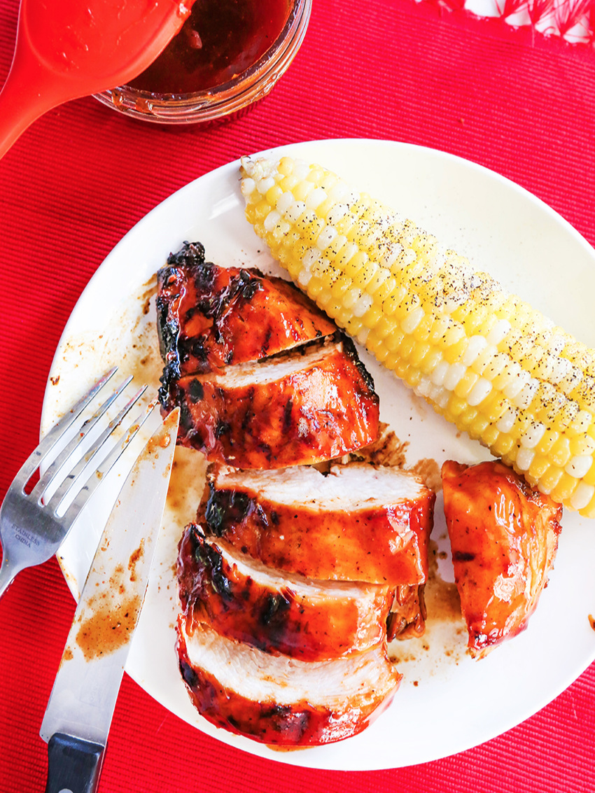 Plate of sliced up grilled chicken and BBQ sauce, corn on the cob on a plate with a fork and knife. 