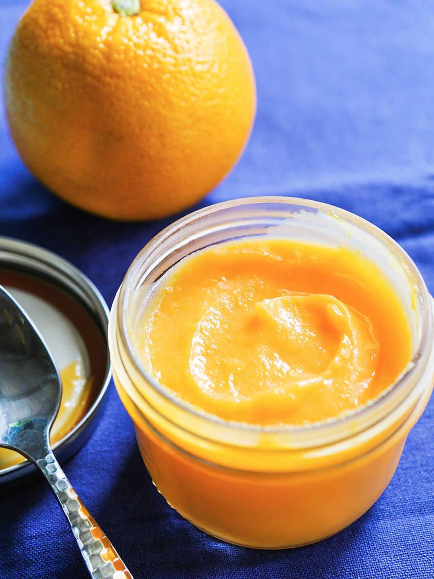 spoon sitting next to a jar of orange curd on a blue tablecloth with a whole orange next to it
