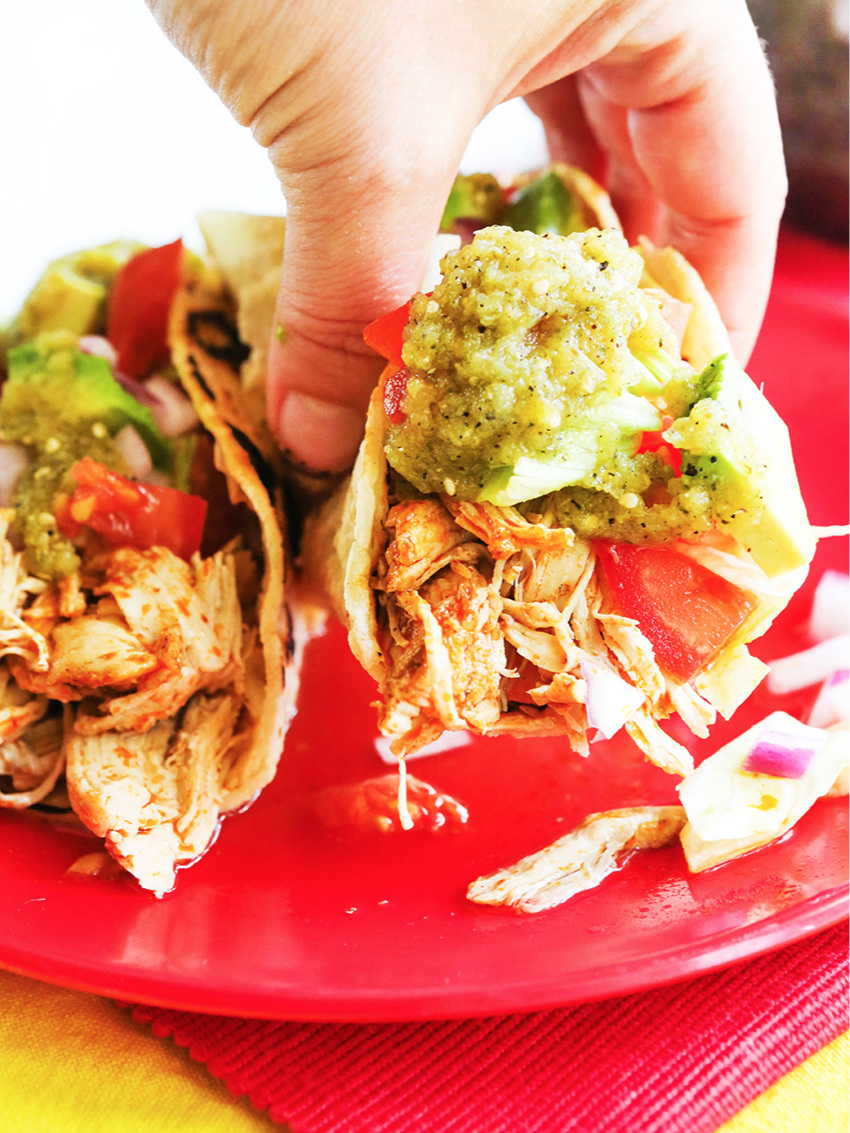 Hand pulling a chicken taco away from plate with green salsa dripping down front.