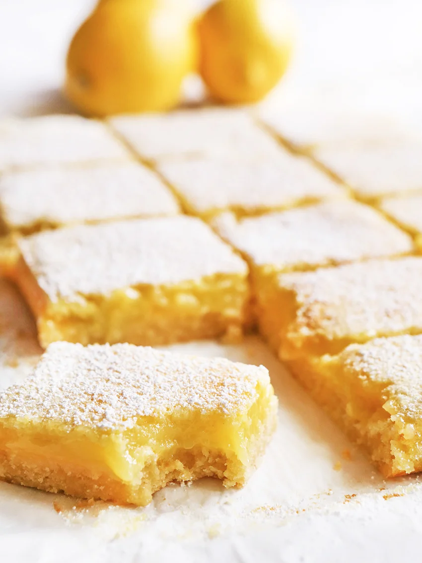 Lemon bars on parchment paper sliced into squares with one bar moved to the front with a bite taken out of it