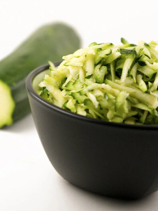  bowl of shredded zucchini next to a zucchini 
