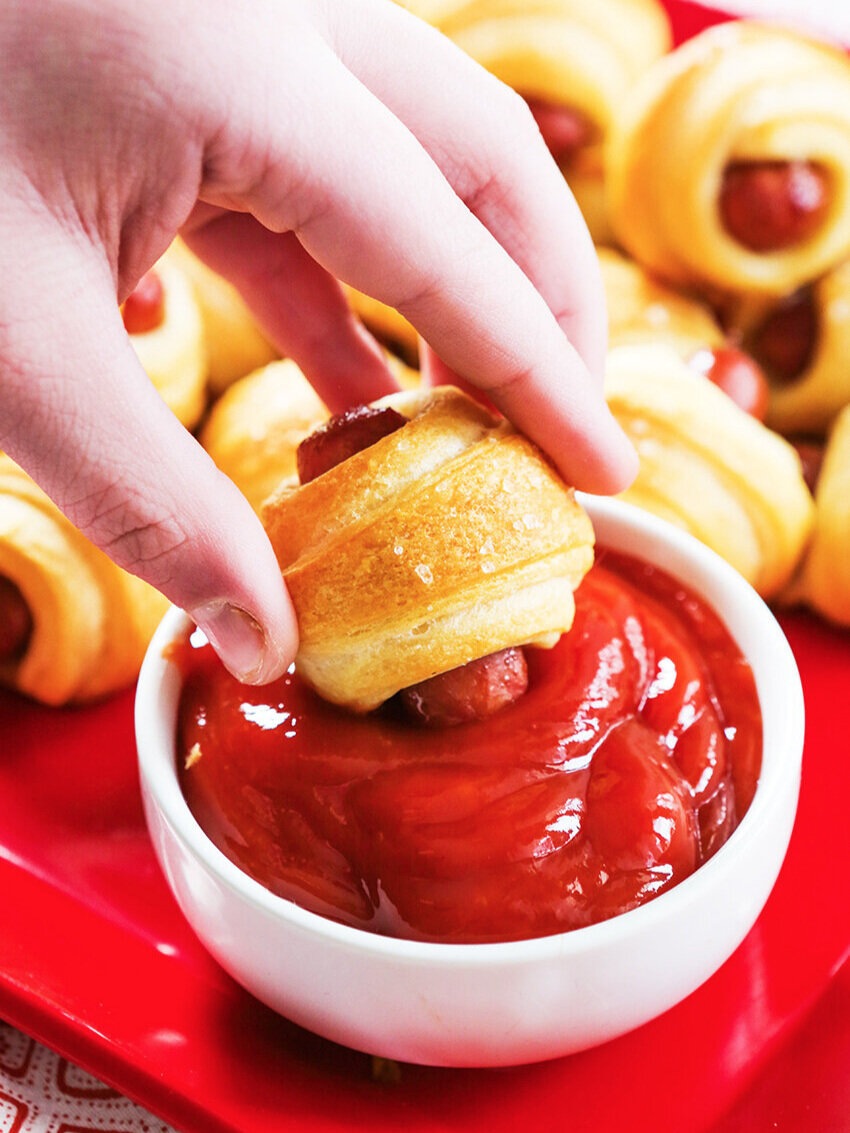 Hand dunking a pig in a blanket into a small bowl of ketchup 