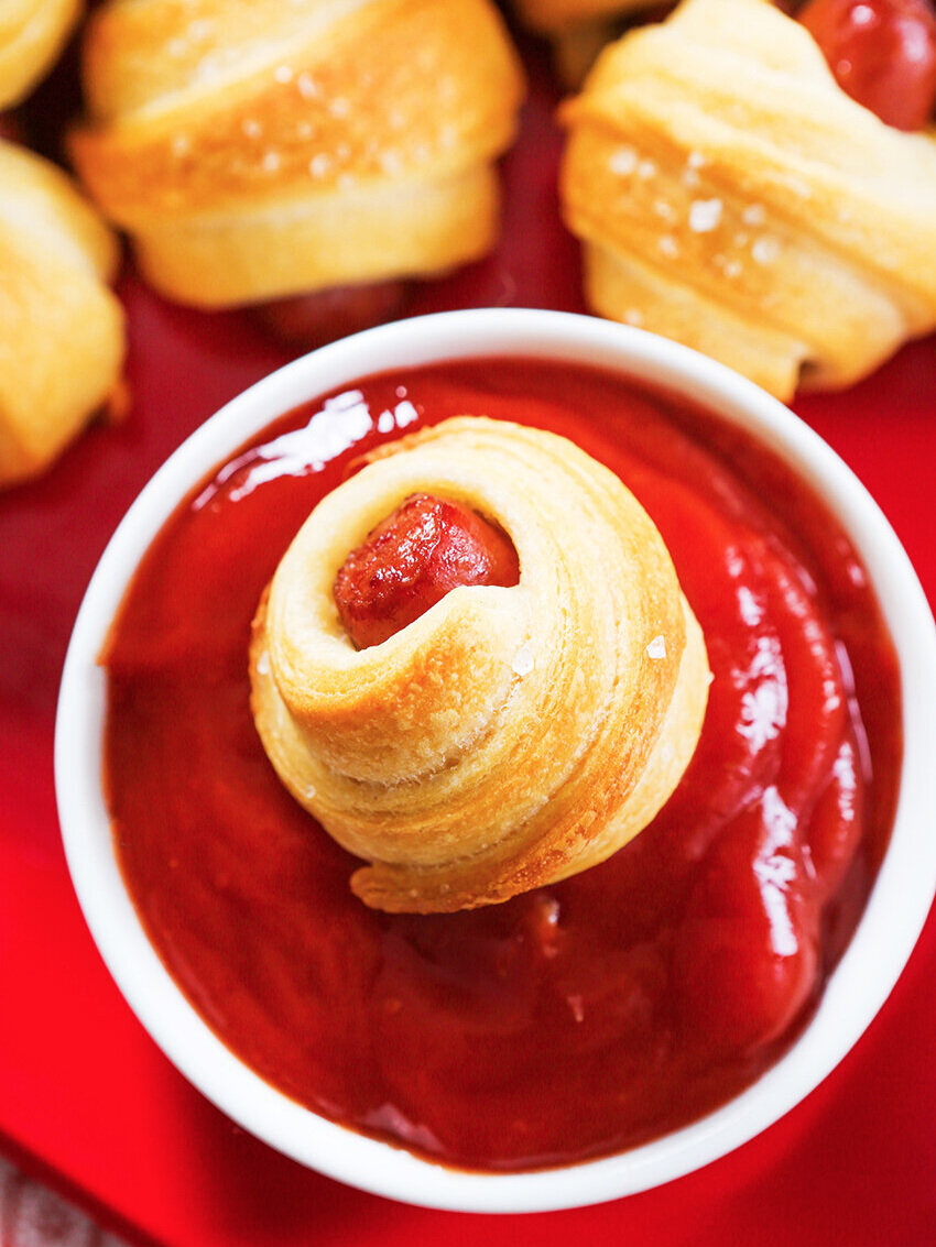 A pig in a blanket dunked into bowl of ketchup 