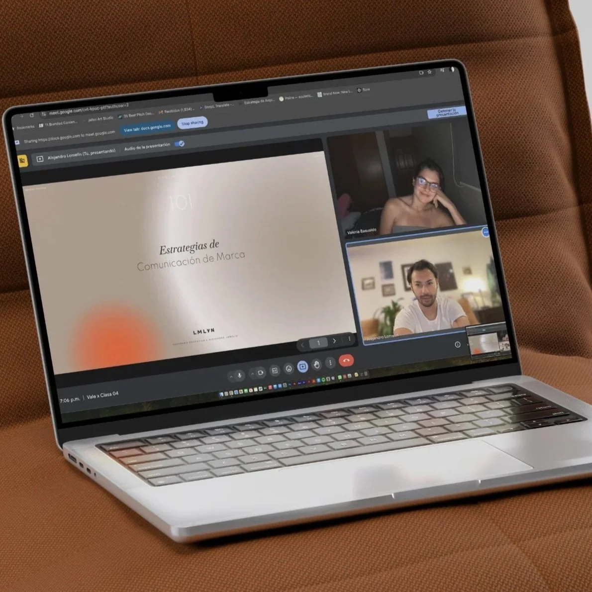 A laptop on a brown couch screen showing a virtual meeting with two people and a presentation slide titled 'Estrategias de Comunicación de Marca'.