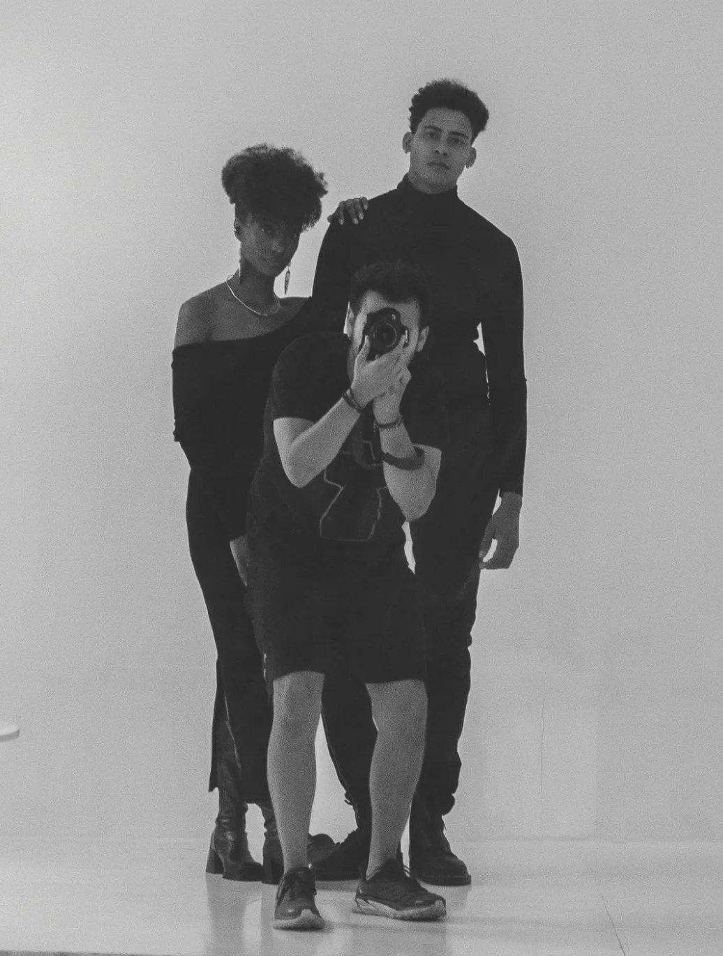 Three people standing in front of a mirror, taking a photo of themselves. The person in the front is holding a camera, the woman on the left with curly hair and off-the-shoulder top, and the man on the right with short curly hair, all dressed in dark clothing.