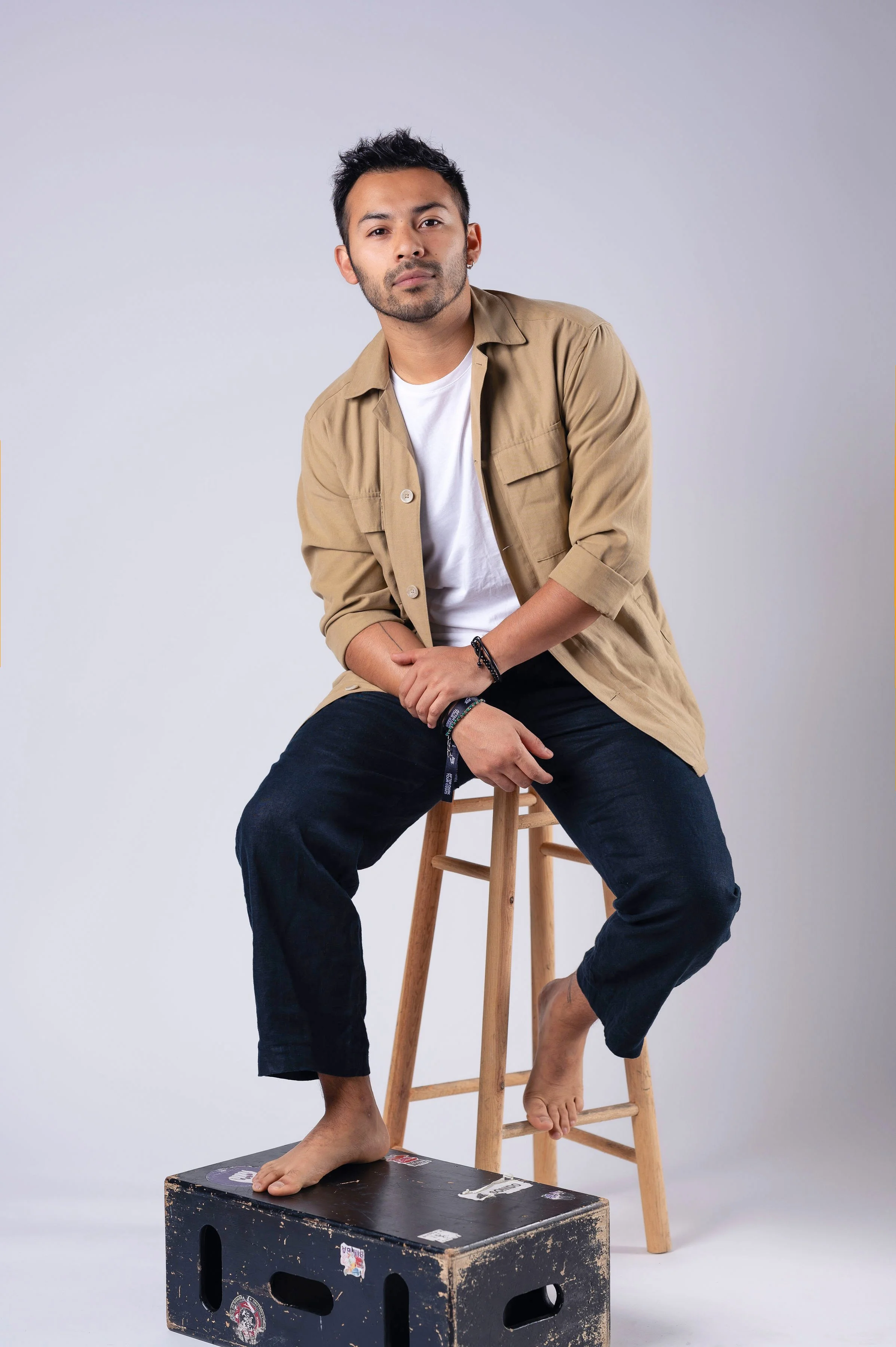 A young man sitting on a wooden stool with one foot on a vintage black box, wearing a beige shirt over a white T-shirt and dark pants, with a neutral expression against a plain white background.
