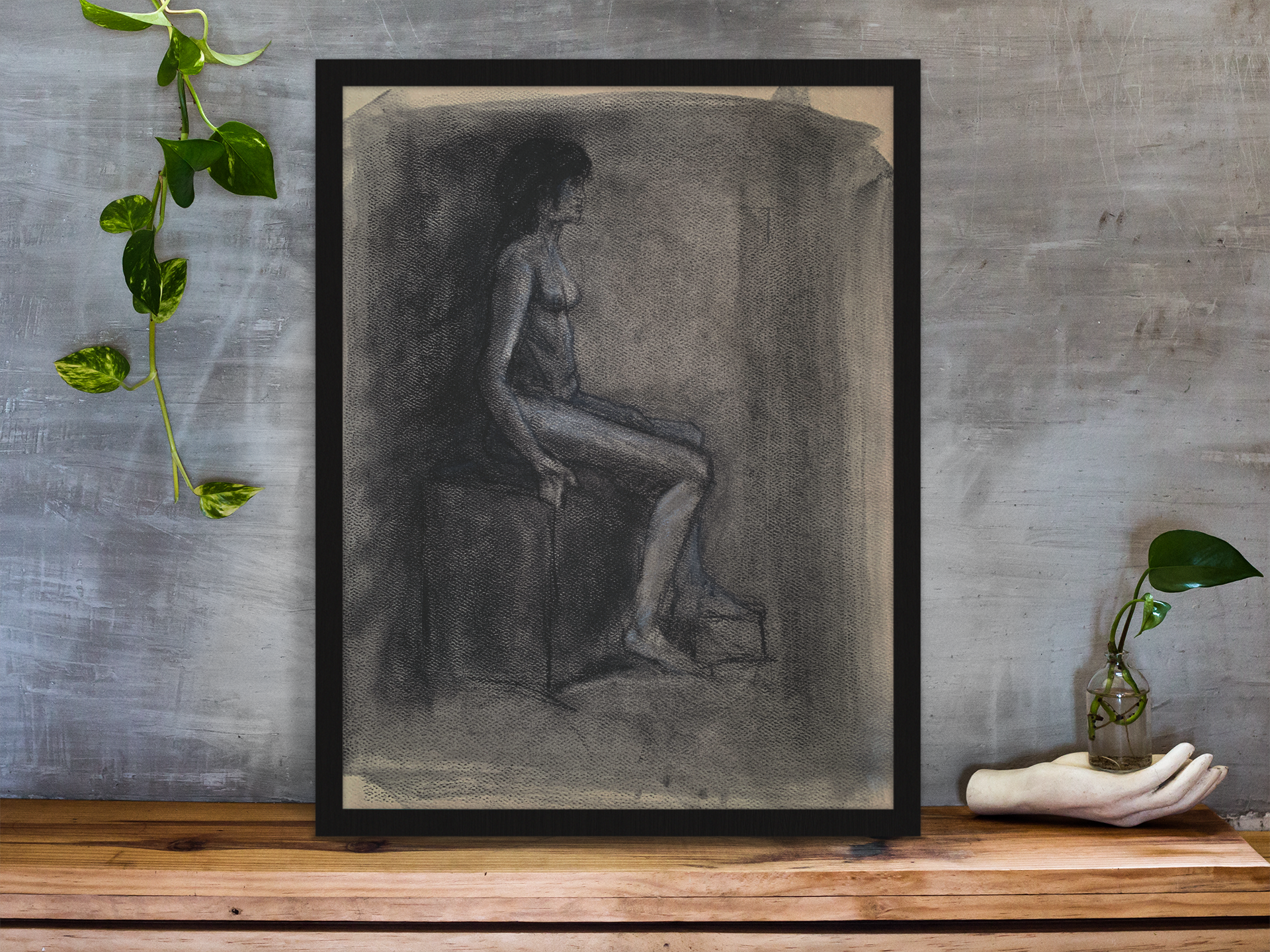 framed-art-print-standing-on-a-wooden-desk-against-a-concrete-wall-near-a-hand-decoration-a10403.png