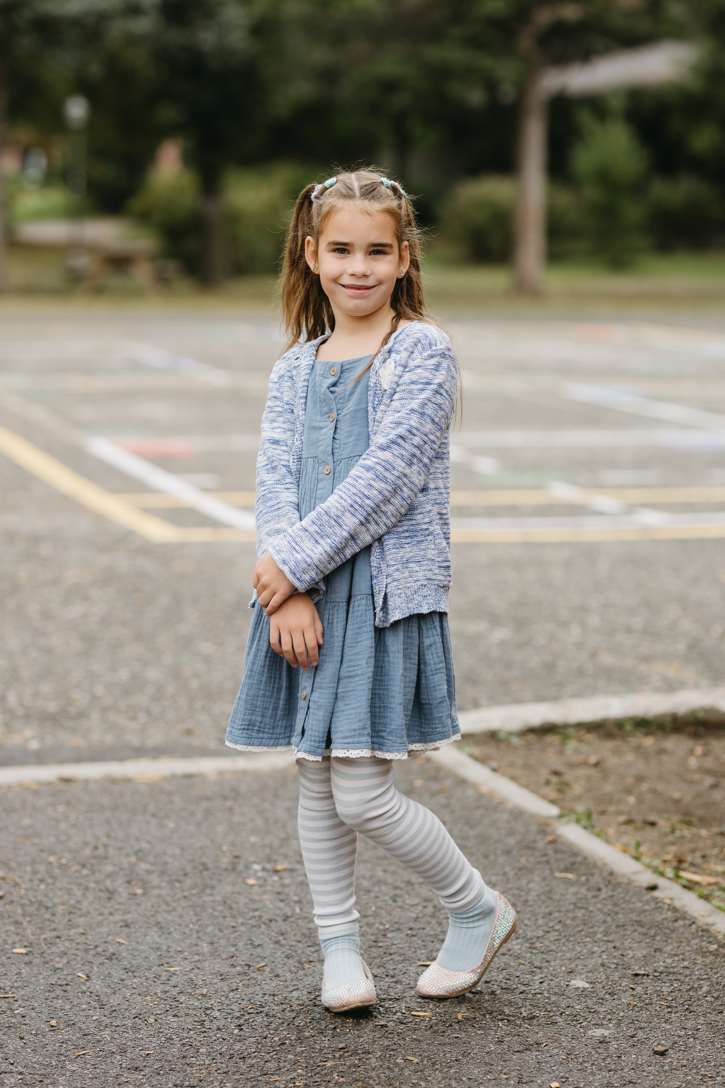 Jeune fille souriante portant une robe bleue, des collants rayés gris et blanc, des chaussures à paillettes, et un cardigan à rayures bleues, dans un parc