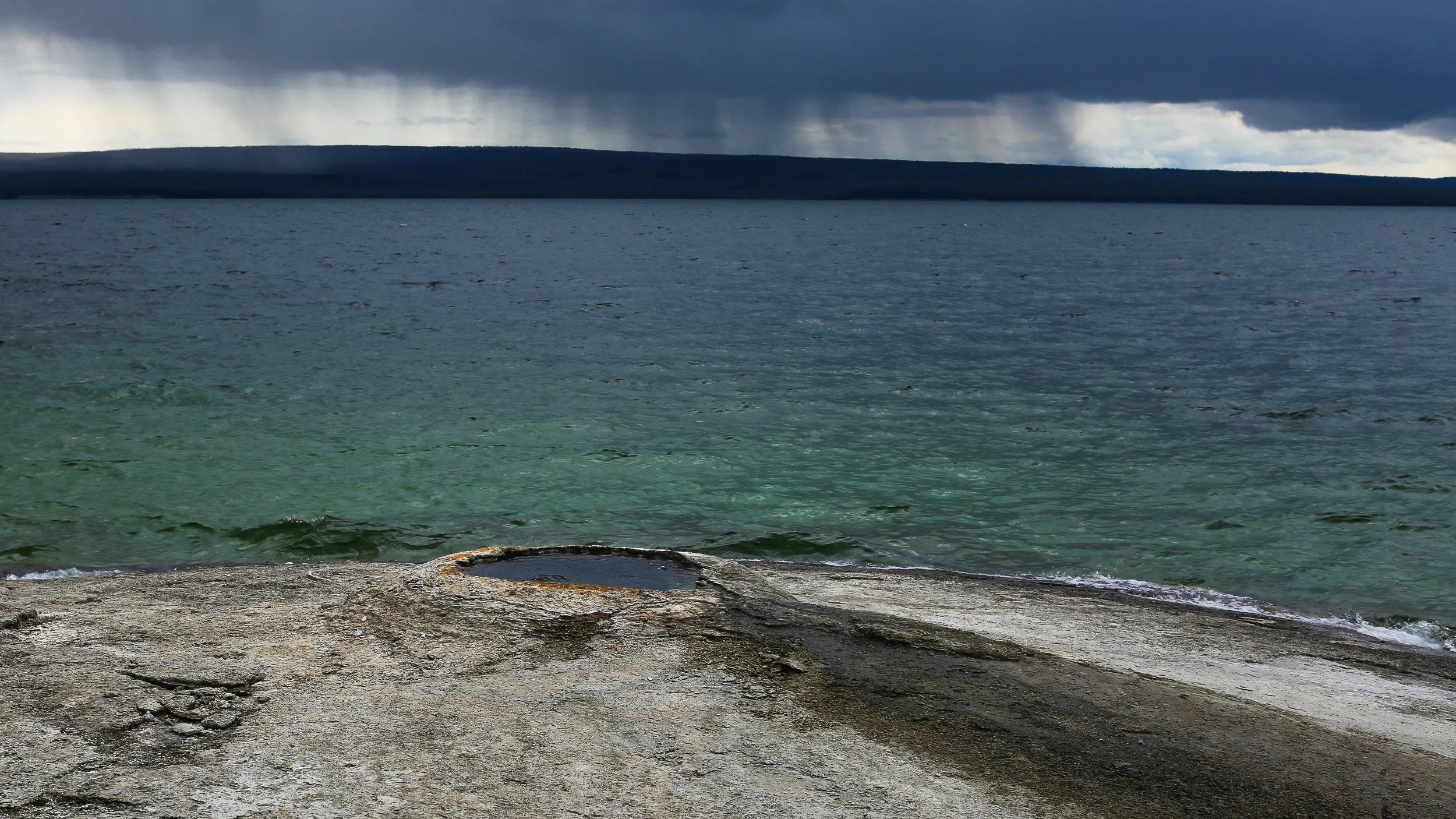 Big Cone, West Thumb Geyser Basin, Yellowstone. Behind the cone is Yellowstone Lake, with rain falling on the Absaroka mountains. Shot taken with a 50mm lens on my Canon 5D, ISO 100, f/9.0, and a longish exposure of 1/50 seconds. "Jutting out into Lake Yellowstone, Big Cone, a companion to Fishing Cone, is a large sinter cone, and is sometimes seen completely submerged by the lake. Eruptions from this geyser are rare, and only reach heights of one foot or less." - West Thumb Interactive Tour