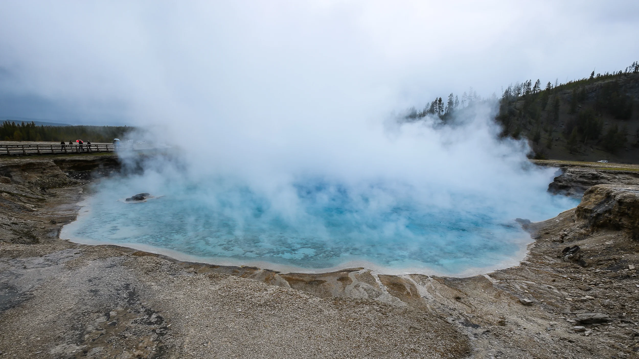 Excelsior Geyser. This geyser is HUGE, and discharges fast-flowing run-off into the adjacent Firehole River. As the hot run-off flows away from the geyser, it cools and allows for colorful gradients (distinct zones) of photosynthetic bacteria and other microbial communities. Shot with ISO 100, f/8.0, 1/160 seconds, wide-angle lens set to 16mm. Temperature: 199°F. Dimensions 276x328 feet. "Excelsior was once the largest geyser In the world. However, the last known major eruptions occurred during the 1880s, when there were numerous eruptions up to 300 feet. The violent eruptions of the 1880s may have caused damage to the siliceous sinter lining, allowing gas leakage and the loss of thermal energy. No observed eruptions were known until 1985 when it erupted for two days. However, it only obtained a height of 20-80 feet. Since its eruptive activity in the 1880s, Excelsior is now a productive thermal spring, presently discharging 4050 gallons per minute. Numerous vents boil and churn the water within the crater, covering it in a dense layer of steam." - yellowstonenationalpark.com