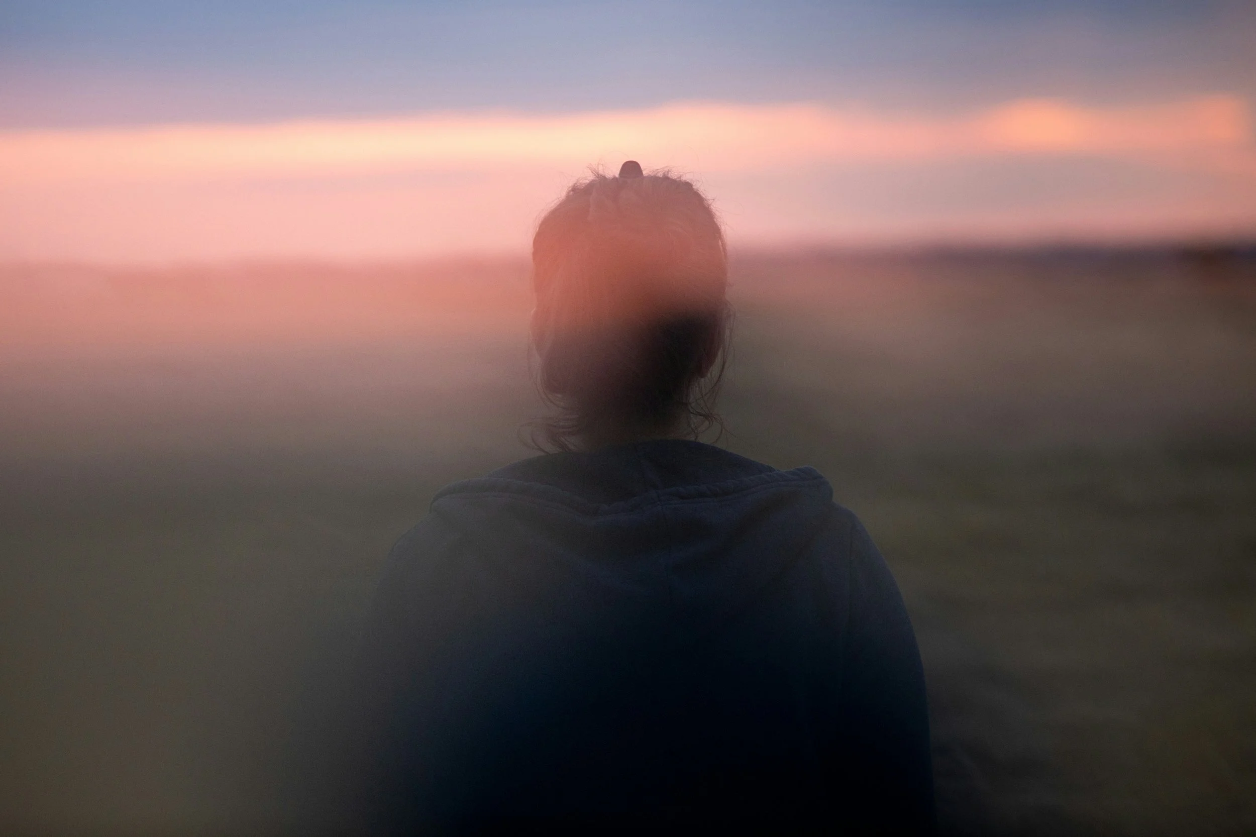A soft, blurred silhouette standing in an open landscape at dusk, partially obscured by mist and fading light, evoking introspection, presence and the quiet distance between self-awareness and direct experience.