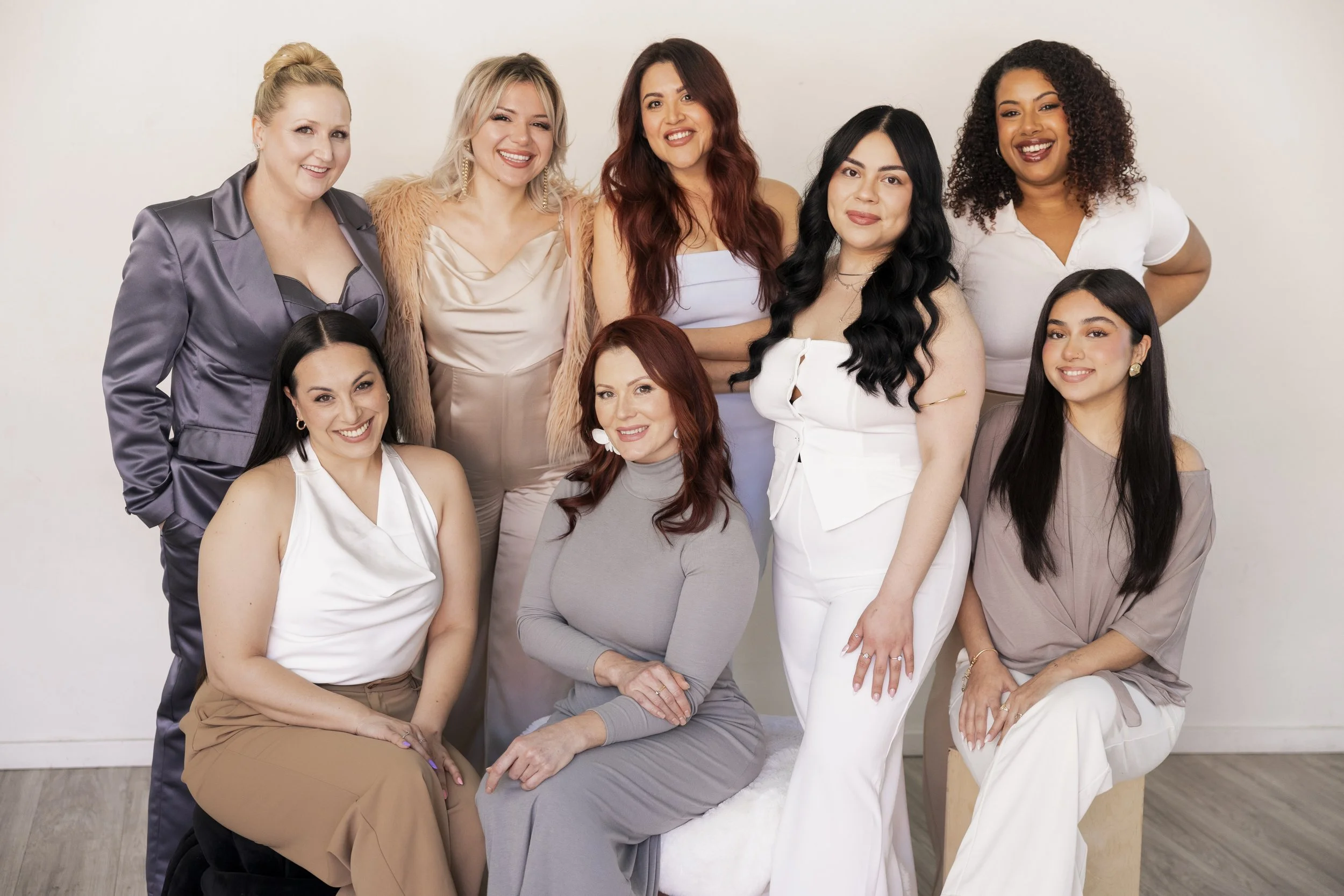 A group of nine diverse women posing together indoors against a plain white wall. Some are sitting, some standing, all smiling and looking at the camera. The women are dressed in a variety of elegant and casual clothing.