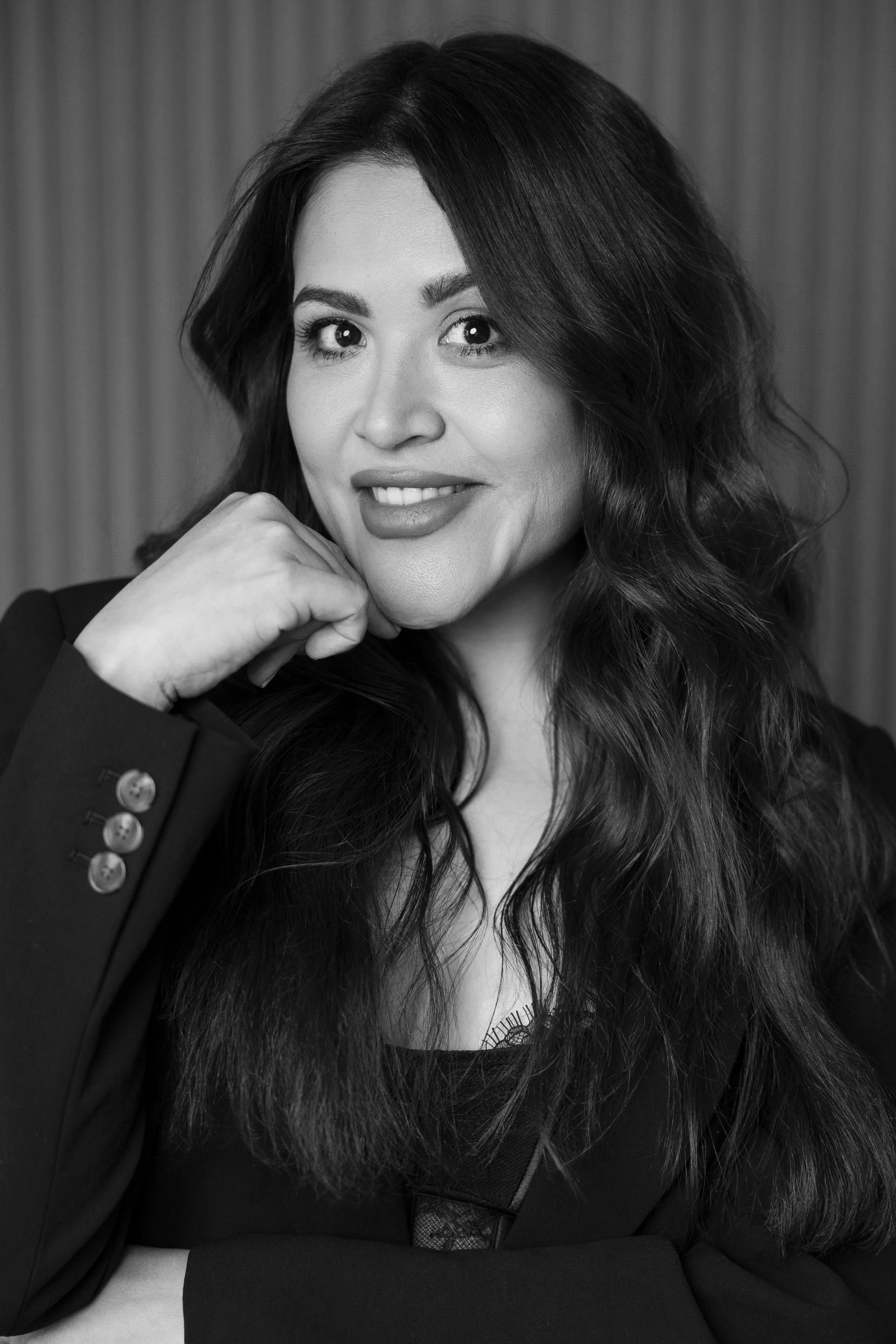 Black and white portrait of a woman with long, wavy hair, smiling slightly, wearing a dark blazer, resting her chin on her hand.