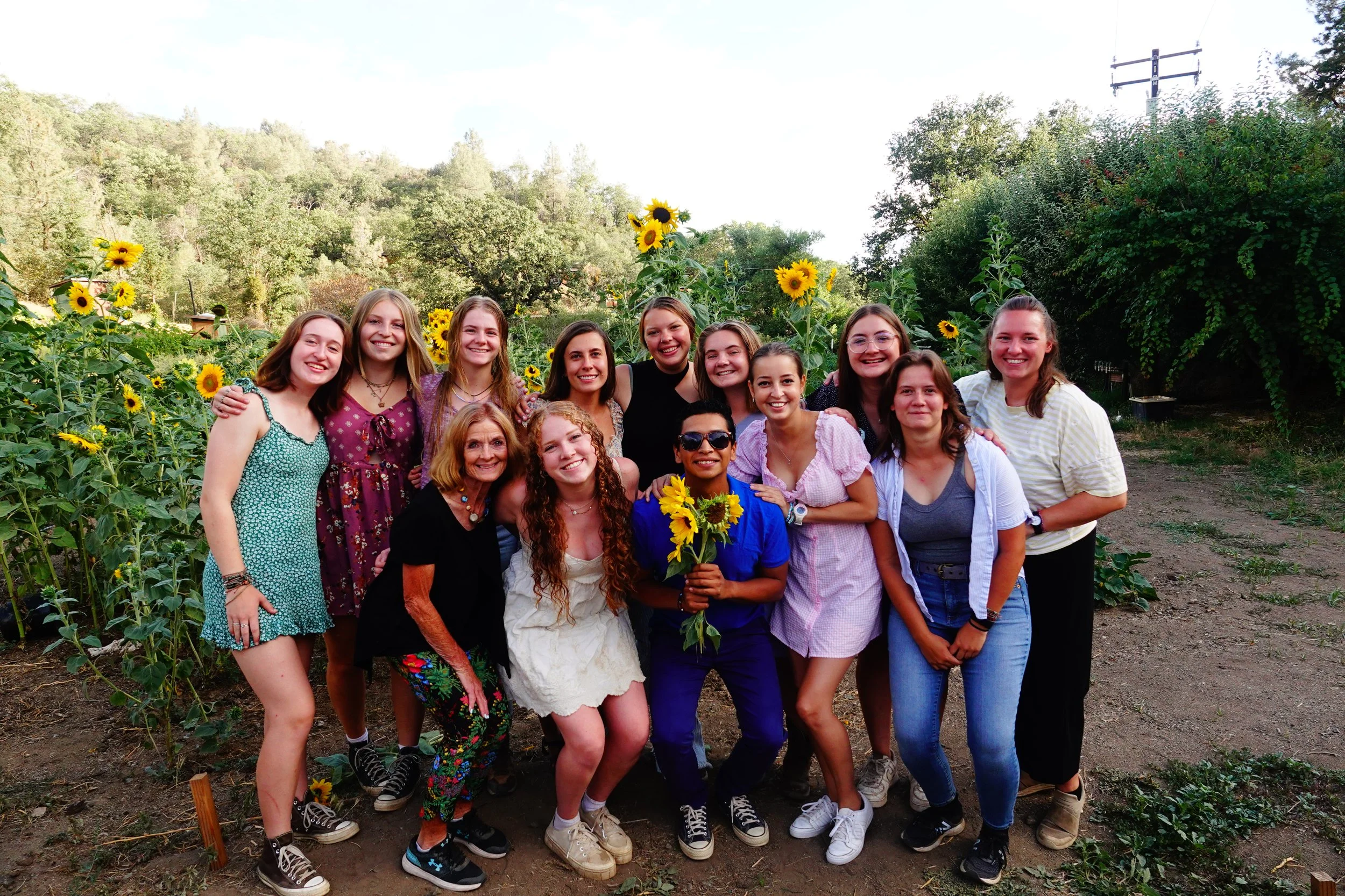 Group of young women and one young man standing together in a sunflower field at Jameson Ranch Camp, smiling at the camera. Bright, sunny day with green trees and hills in the background.