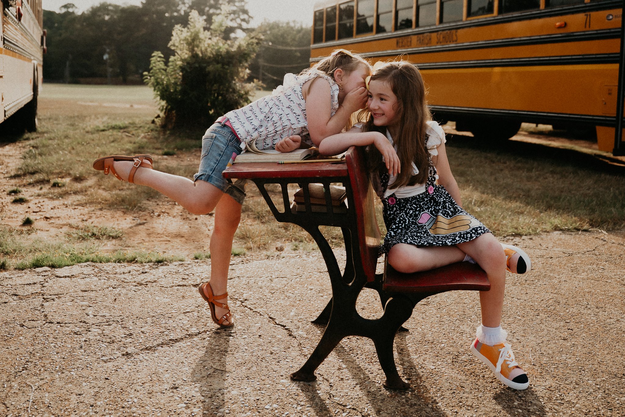 two sisters whispering secrets at a vintage desk between two yellow school busses in back to school clothes featuring pencils and composition books