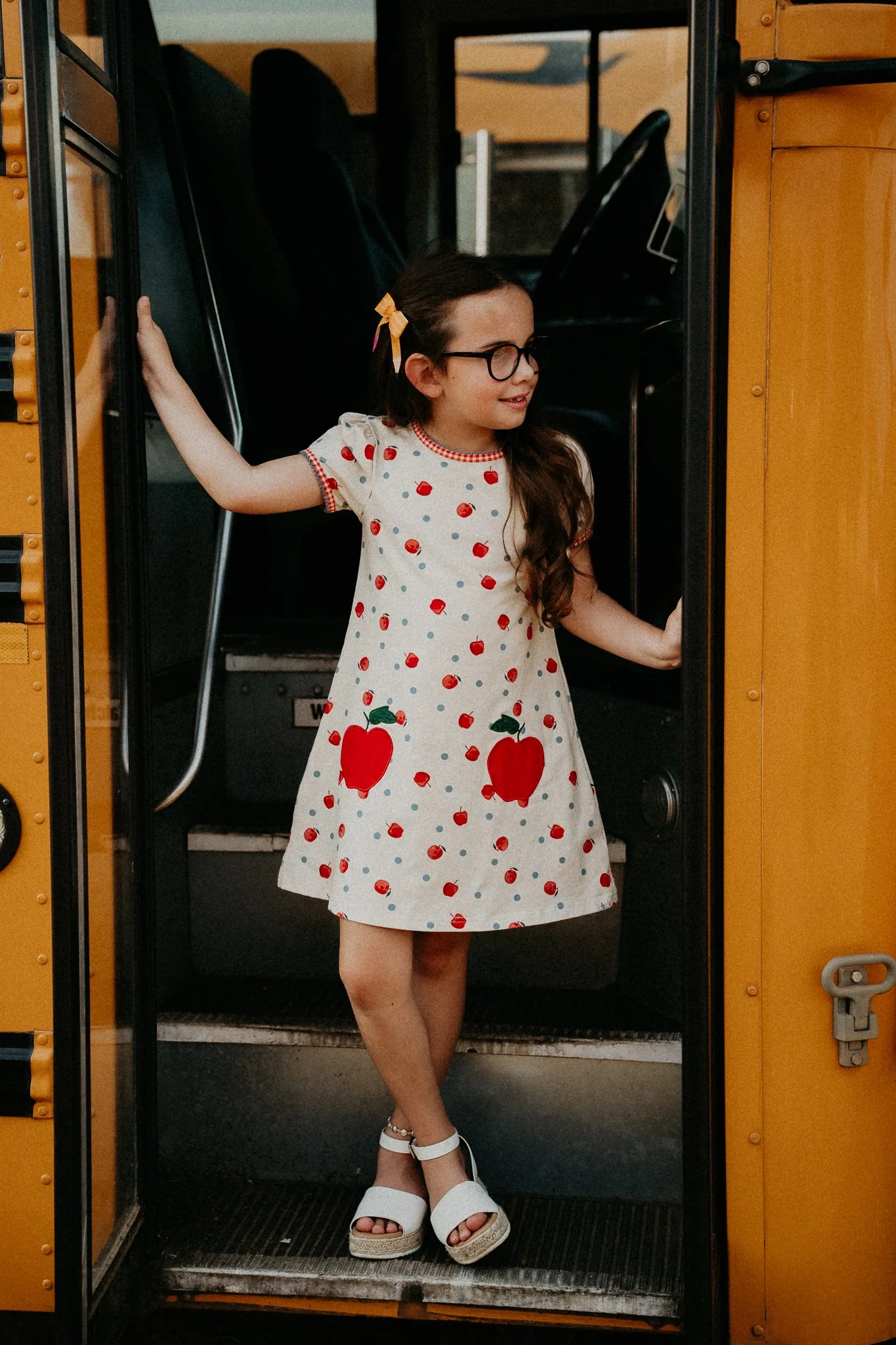 dark haired girl in apple print dress with pencil bow in doorway of vintage yellow school bus looking off into the distance