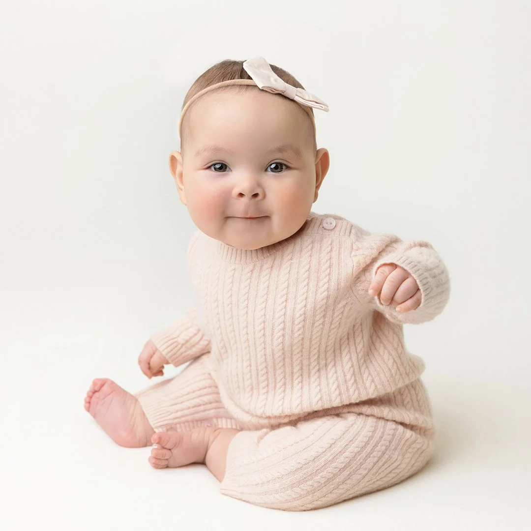 A baby girl in a pink sweater sitting up on an ivory backdrop