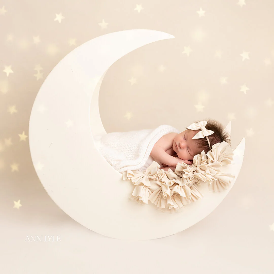 A sleeping baby with a bow headband lying on a ruffled blanket inside a crescent moon prop, against a starry background.