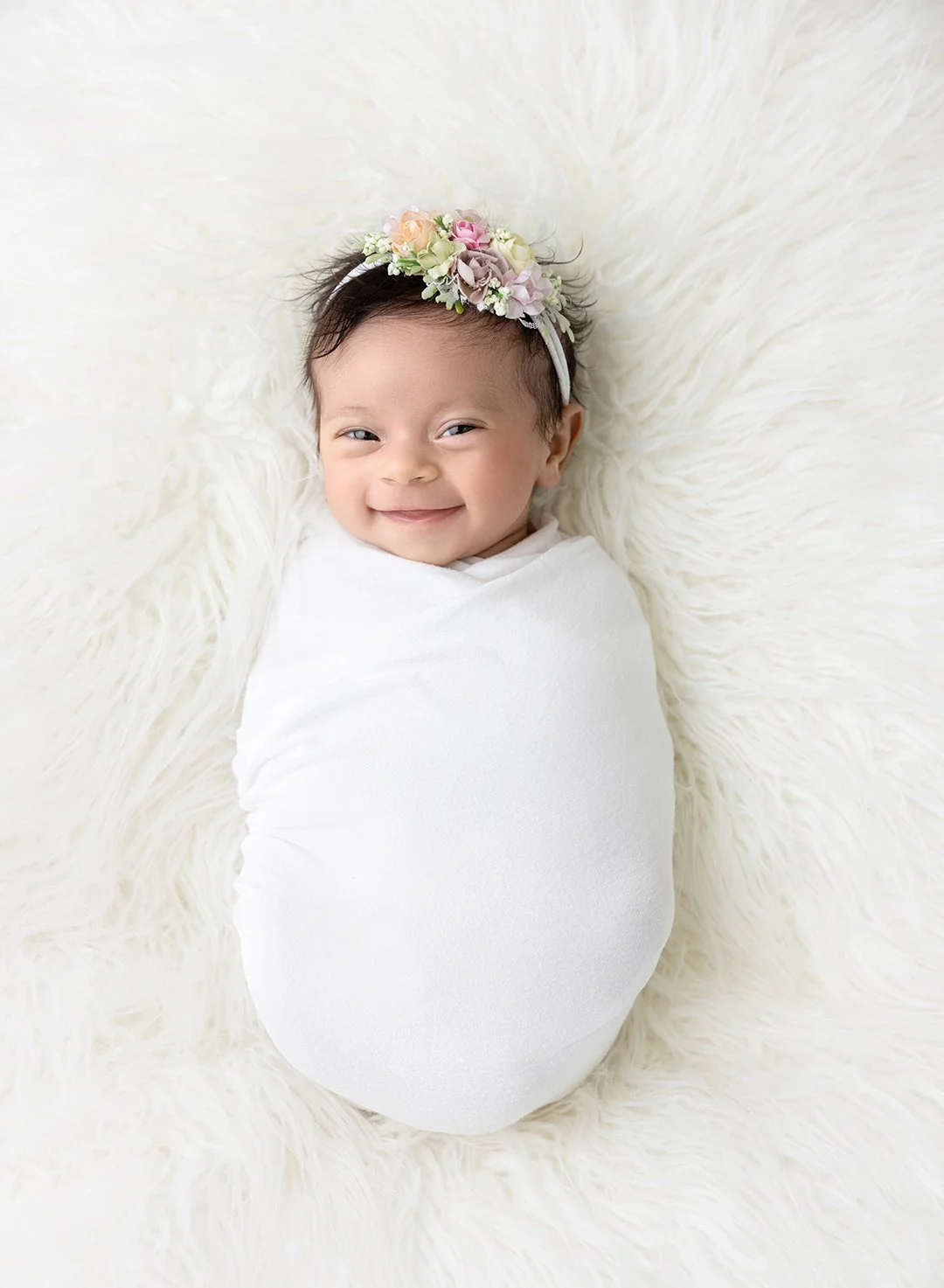 a smiling young baby in a swaddle with a floral headband.