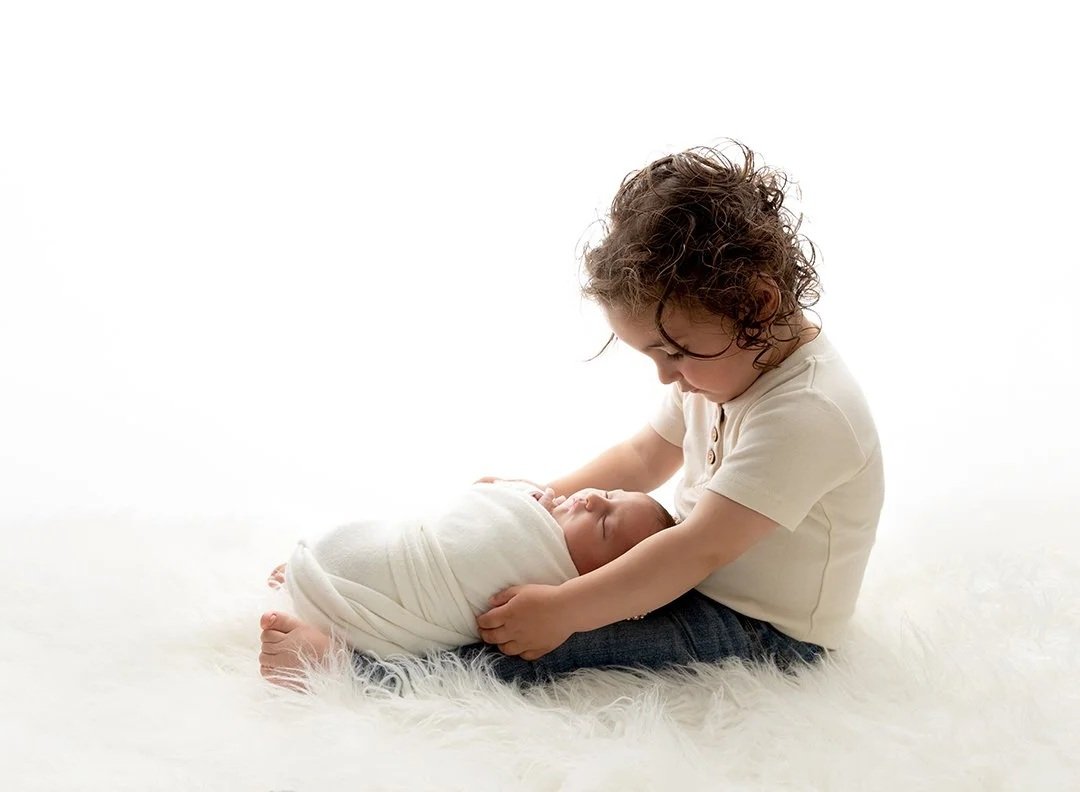 A toddler holding a newborn baby with an all white backdrop