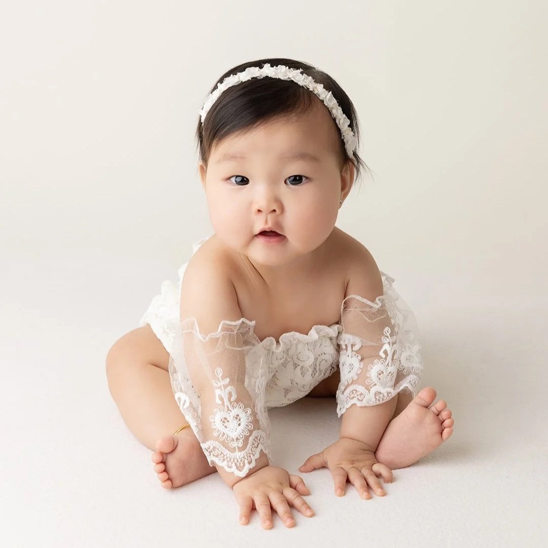 A baby girl with a white dress sitting up looking at the camera
