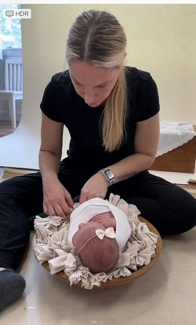 A woman sitting cross-legged looks down at a newborn baby lying in a bassinet, wrapped in a white blanket, with a white hat on the baby's head.