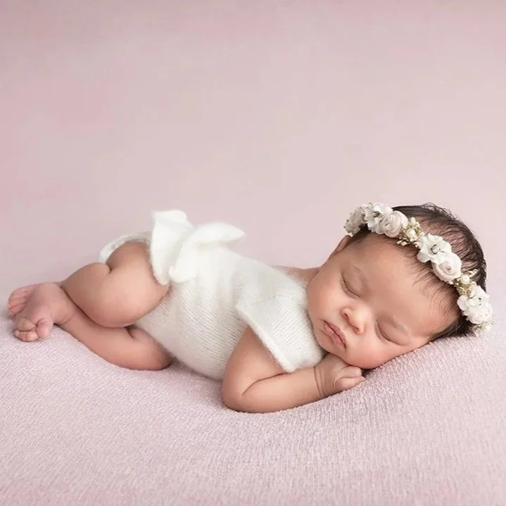 A sleeping baby girl lying on her stomach on a pink blanket, wearing a white dress and a flower headband.